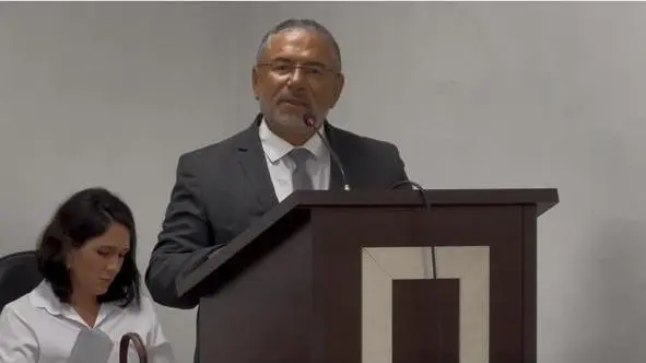 Juiz José Antônio Bezerra Filho usa terno cinza e óculos, fala ao microfone em um púlpito de madeira escura. Ao lado esquerdo inferior, uma mulher de camisa branca olha para baixo, sentada em uma mesa.