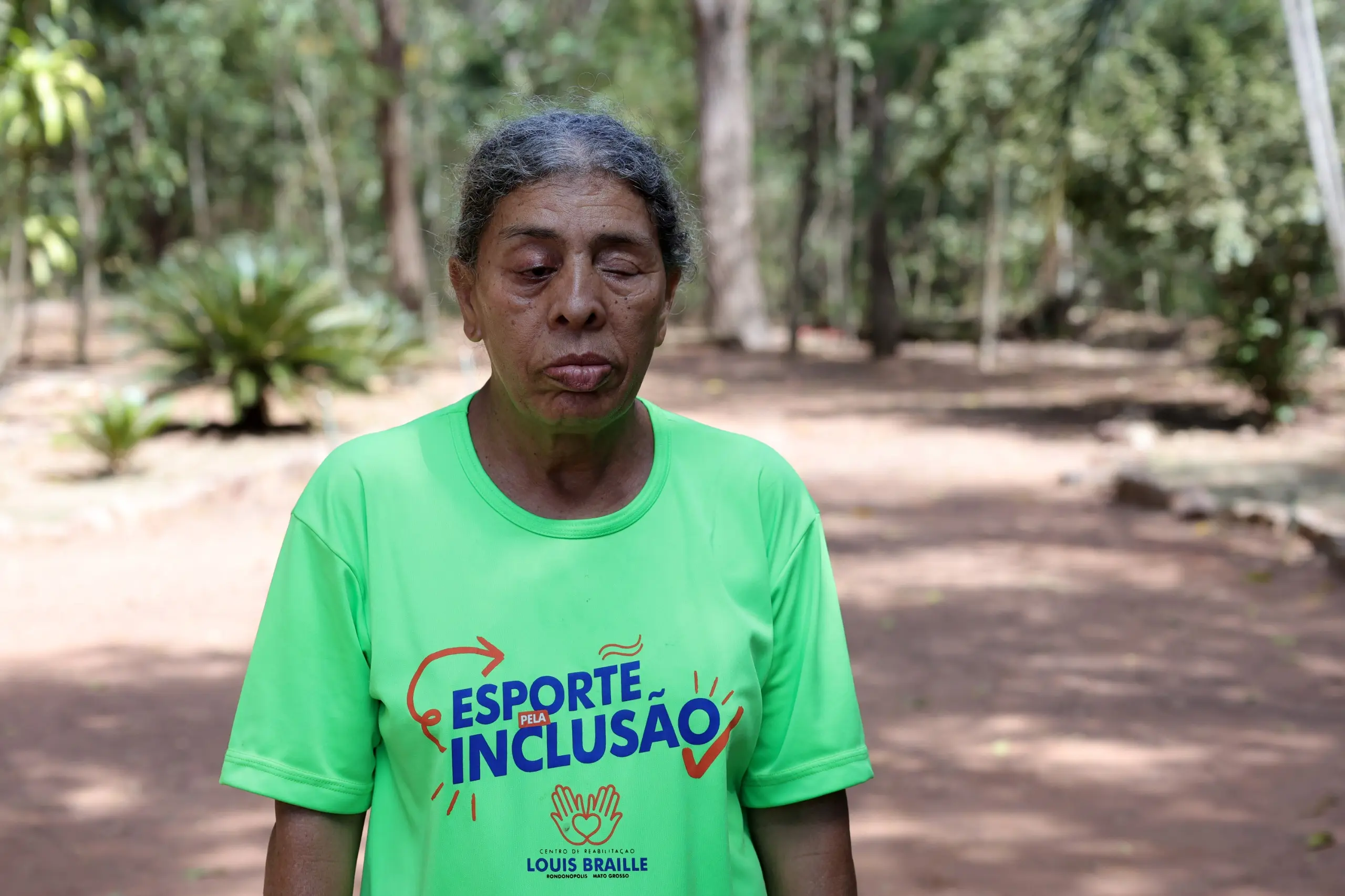 Mulher negra de cabelos grisalhos presos, vestindo camiseta verde com a inscrição Esporte pela Inclusão. Ela está em pé em um caminho de terra cercado por árvores e vegetação.