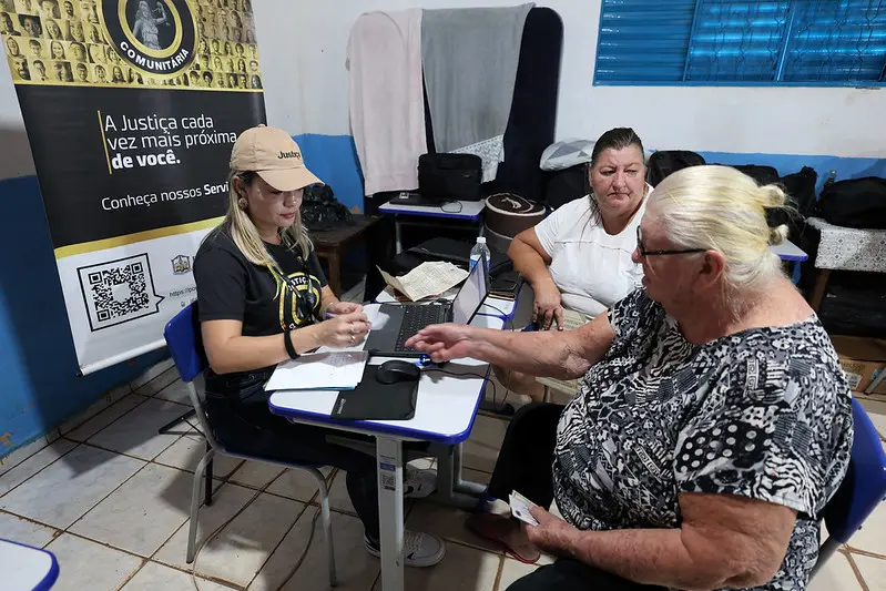 Foto horizontal colorida, que mostra a assessora jurídica Juliana de Paula, atendendo Roseli e Iraci, durante mutirão Justiça em Ação, numa sala de aula. Ela é uma mulher branca, loira, usando boné bege e camiseta preta da Justiça Comunitária. Atrás dela, há um banner da Justiça Comunitária.