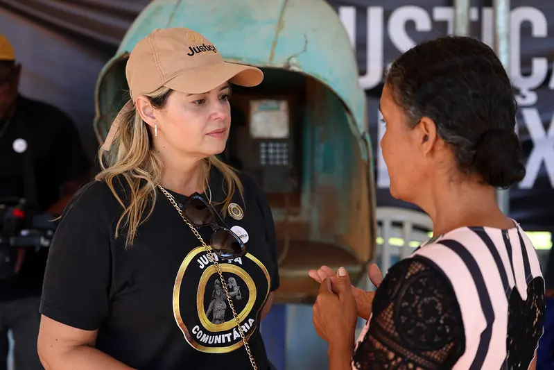 Foto horizontal colorida, em plano médio, que mostra a assessora da Justiça Comunitária Ana Carla Bock conversando em pé com uma mulher, durante mutirão Justiça em Ação. A assessora é uma mulher branca, loira, usando boné bege da Justiça em Ação e camiseta preta da Justiça Comunitária. 