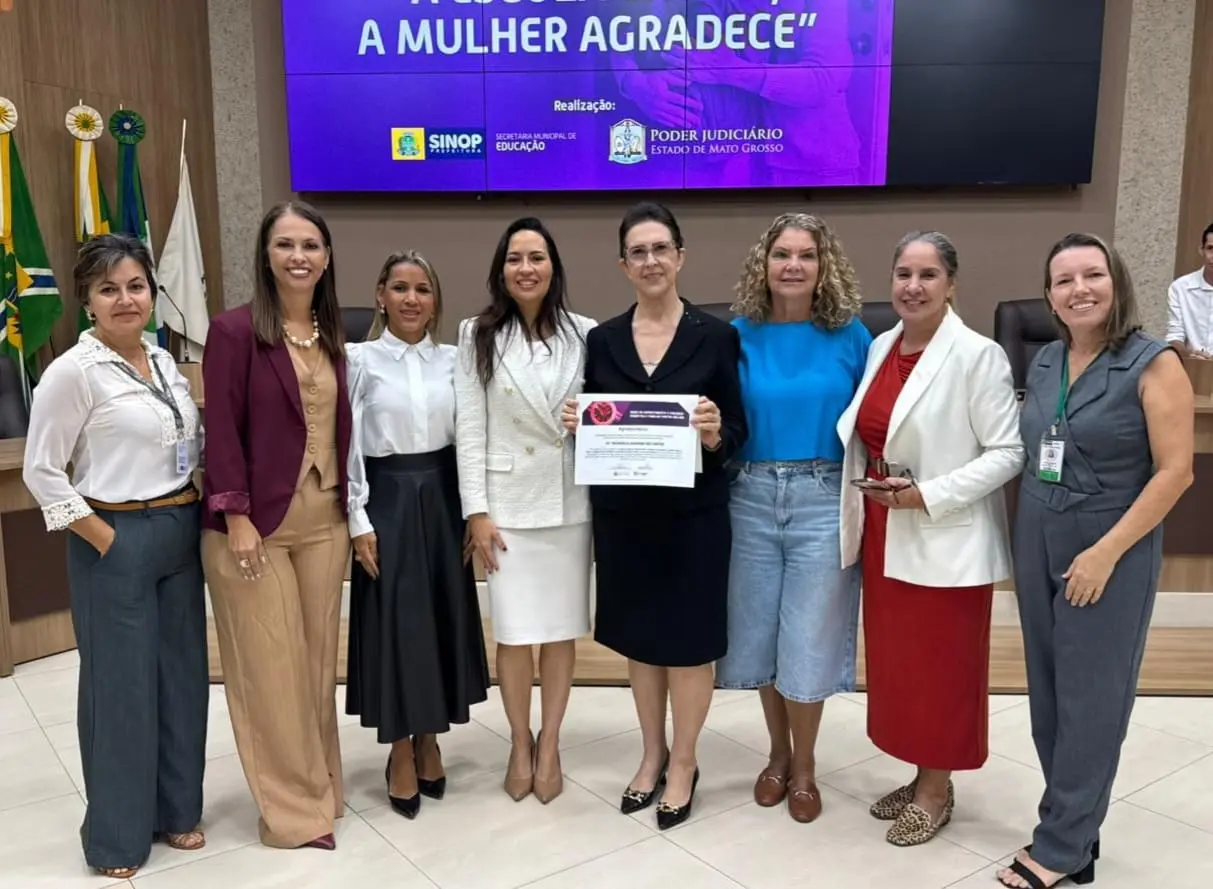 Oito mulheres posam alinhadas em um auditório. A mulher ao centro, de vestido preto, segura um certificado impresso. Atrás delas, um telão roxo exibe logos institucionais.