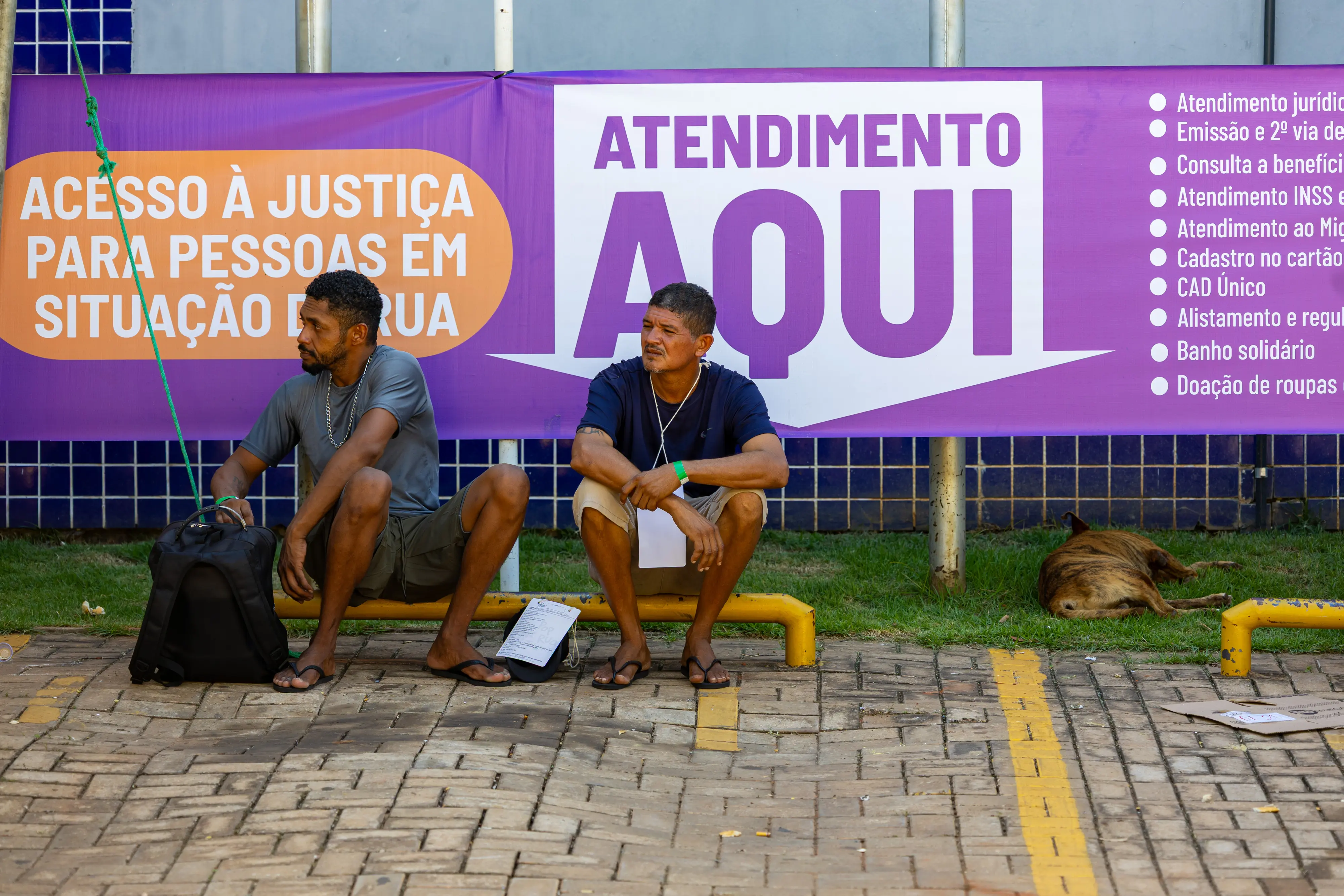 Dois homens sentados em uma mureta baixa diante de banner roxo com a frase 