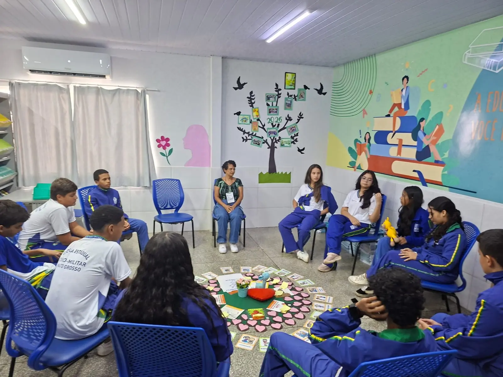 Jovens com uniforme escolar azul e branco estão sentados em círculo de cadeiras em uma sala de aula decorada. No centro do chão, há um arranjo com corações, cartões e objetos simbólicos.