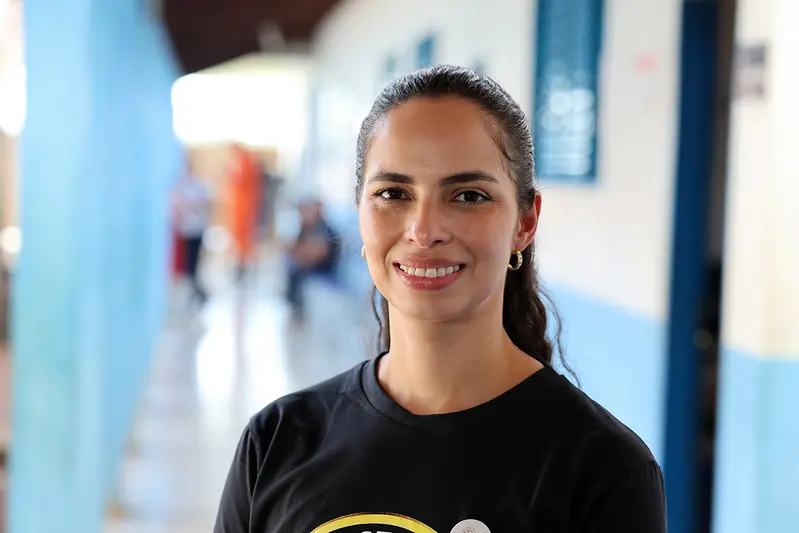 Foto horizontal colorida que mostra a juíza diretora do foro de Paranatinga, Raiane Arteman, posando sorrindo para a foto, no corredor da escola onde ocorreu o mutirão Justiça em Ação. Ela é uma mulher branca, de olhos e cabelos castanhos, usando camiseta preta da Justiça Comunitária.