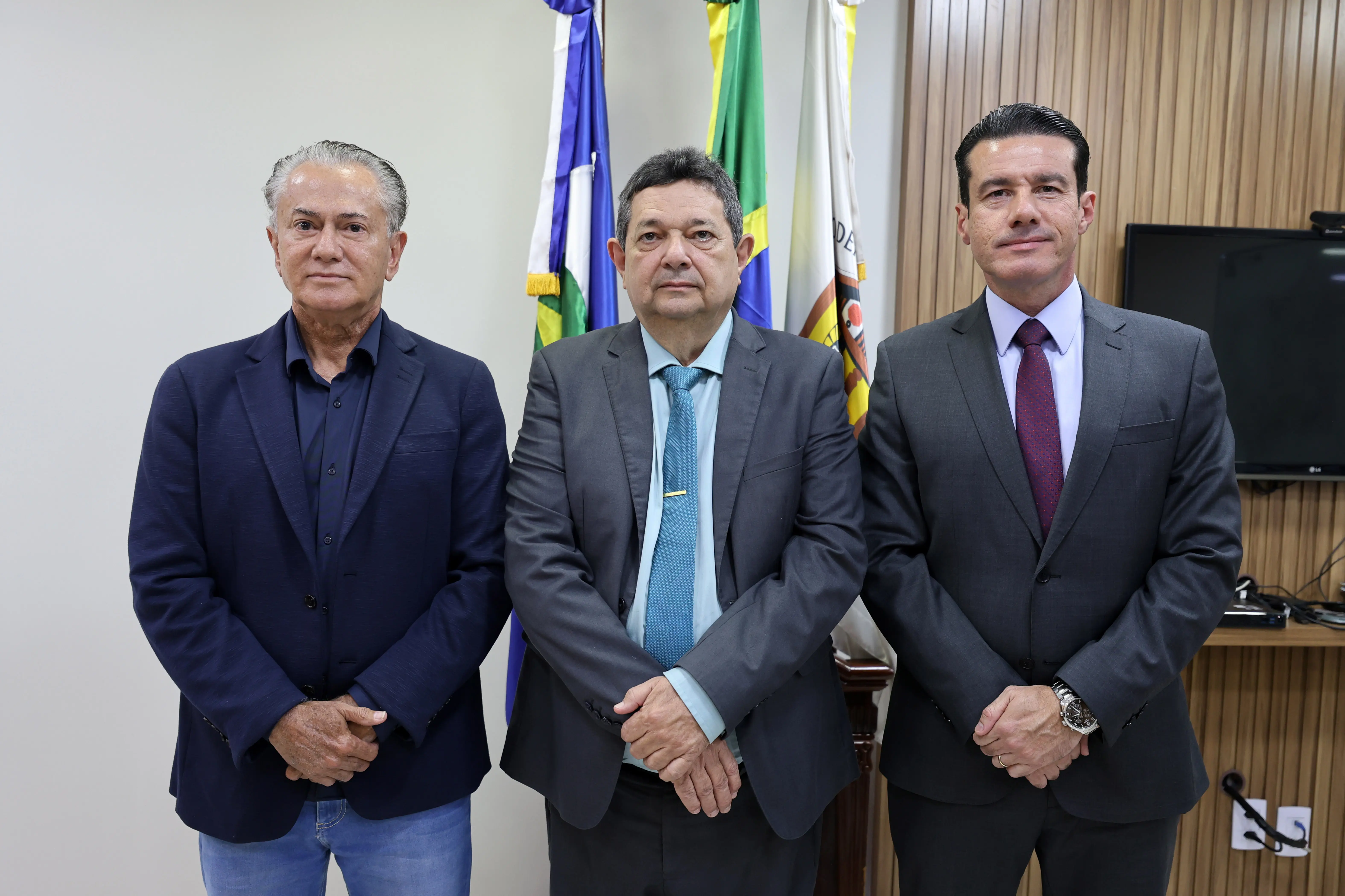 Três homens em trajes formais posam lado a lado em frente a bandeiras oficiais. À esquerda, blazer azul; ao centro, terno cinza com gravata azul; à direita, terno cinza com gravata bordô.