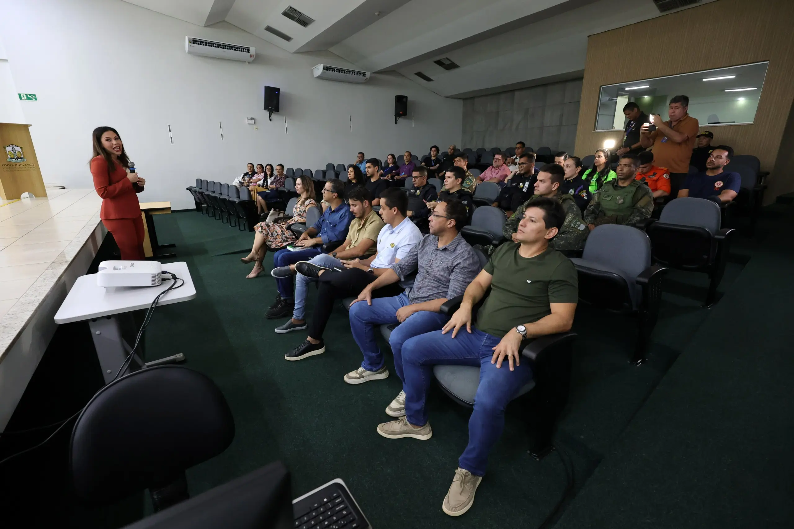 Visão ampla de um auditório com várias pessoas sentadas assistindo a uma palestra. À esquerda, a mulher de terno vermelho fala ao microfone no palco. Há policiais e civis na plateia.