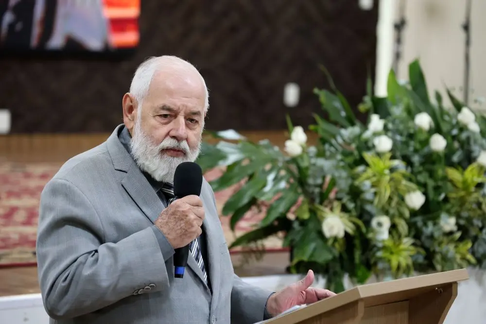 Foto horizontal que mostra o presidente do TJMT, José Zuquim Nogueira, falando ao microfone, no púlpito do auditório. Ele é um senhor branco, de cabelos e barbas brancos, olhos azuis, usando terno cinza. Atrás dele, há um enorme arranjo de flores brancas com folhagens.