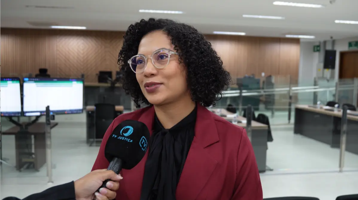 Mulher de cabelos cacheados pretos, óculos transparentes e blazer vinho sobre blusa preta com laço, concede entrevista a microfone da TV Justiça. Ao fundo, sala com monitores.
