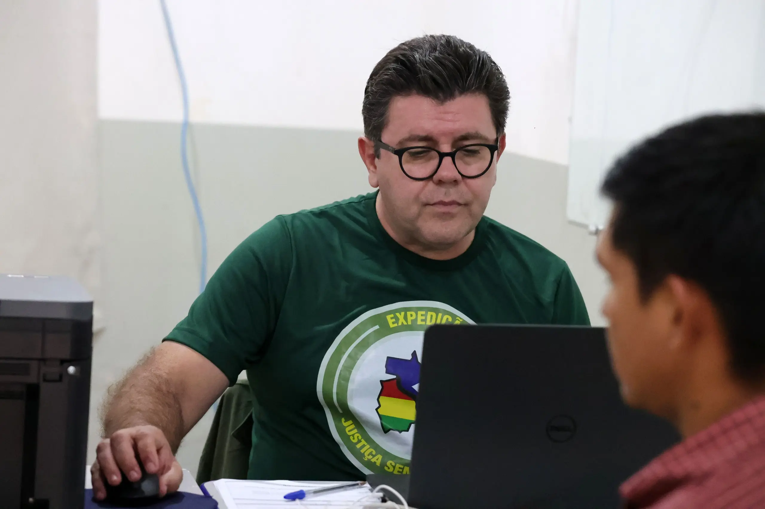 Foto horizontal que mostra o oficial de Registro Civil de Vila Bela da Santíssima Trindade, Ademir Baldo, trabalhando, sentado em frente a um laptop. Ele é um homem branco, de cabelo castanho e liso, usando camiseta verde com logo da Expedição Justiça Sem Fronteiras e óculos de grau.