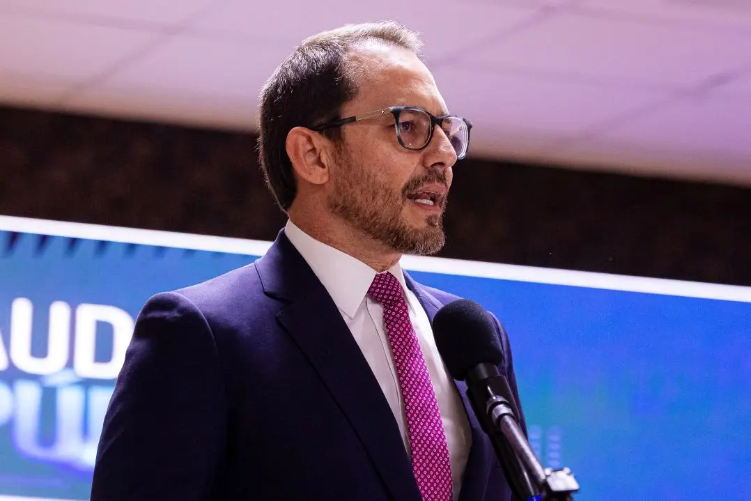 Foto horizontal que mostra o advogado Giovane Santin do busto pra cima, falando ao microfone. Ele é um homem branco, de barba e cabelos castanhos claros, usando camisa branca, gravata púrpura e terno azul marinho e óculos de grau.