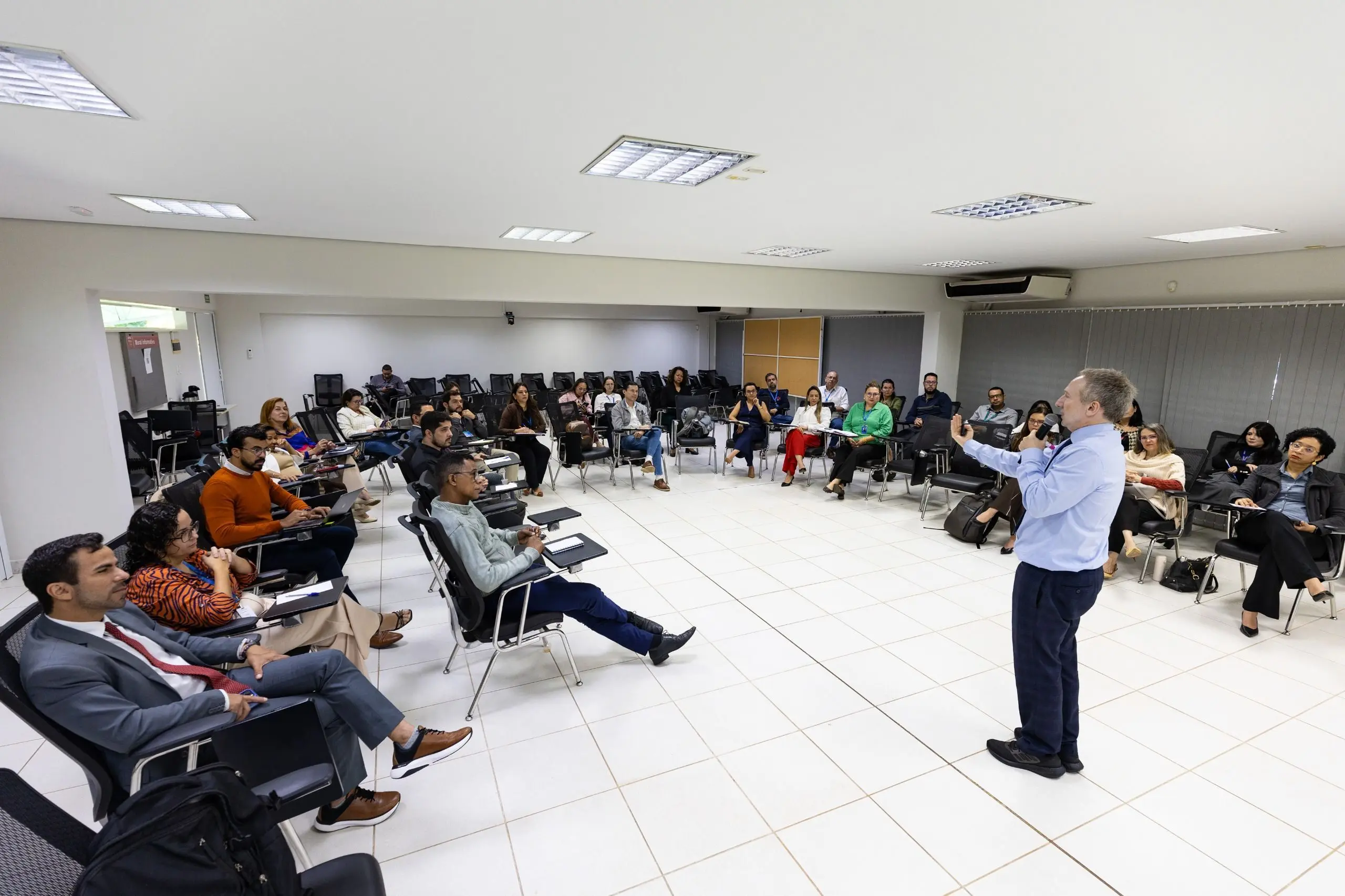 Professor Vlademir gesticula enquanto ministra aula em uma sala com alunos. Os alunos estão sentados em meia lua e prestam atenção ao professor.