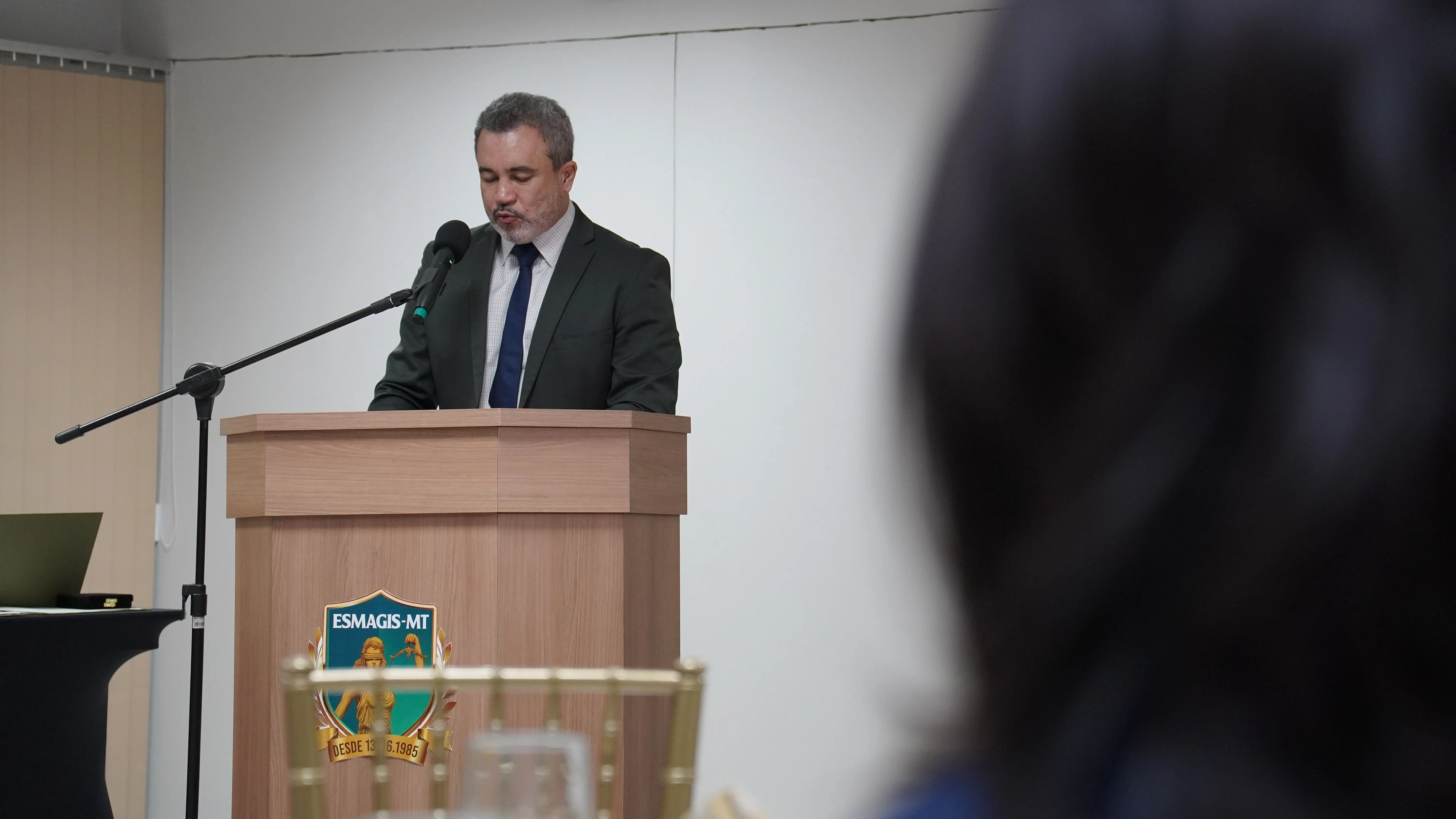 Homem de terno escuro e gravata azul discursa em um púlpito de madeira com o brasão da Esmagis-MT fixado à frente. À direita e em primeiro plano, vê-se a silhueta desfocada da cabeça de alguém.