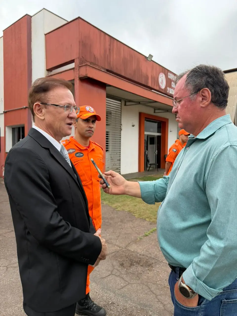 Homem de terno preto dá entrevista a repórter de camisa verde, que segura um celular. Ao fundo, um bombeiro de uniforme laranja observa a cena em frente ao batalhão, sob céu nublado.