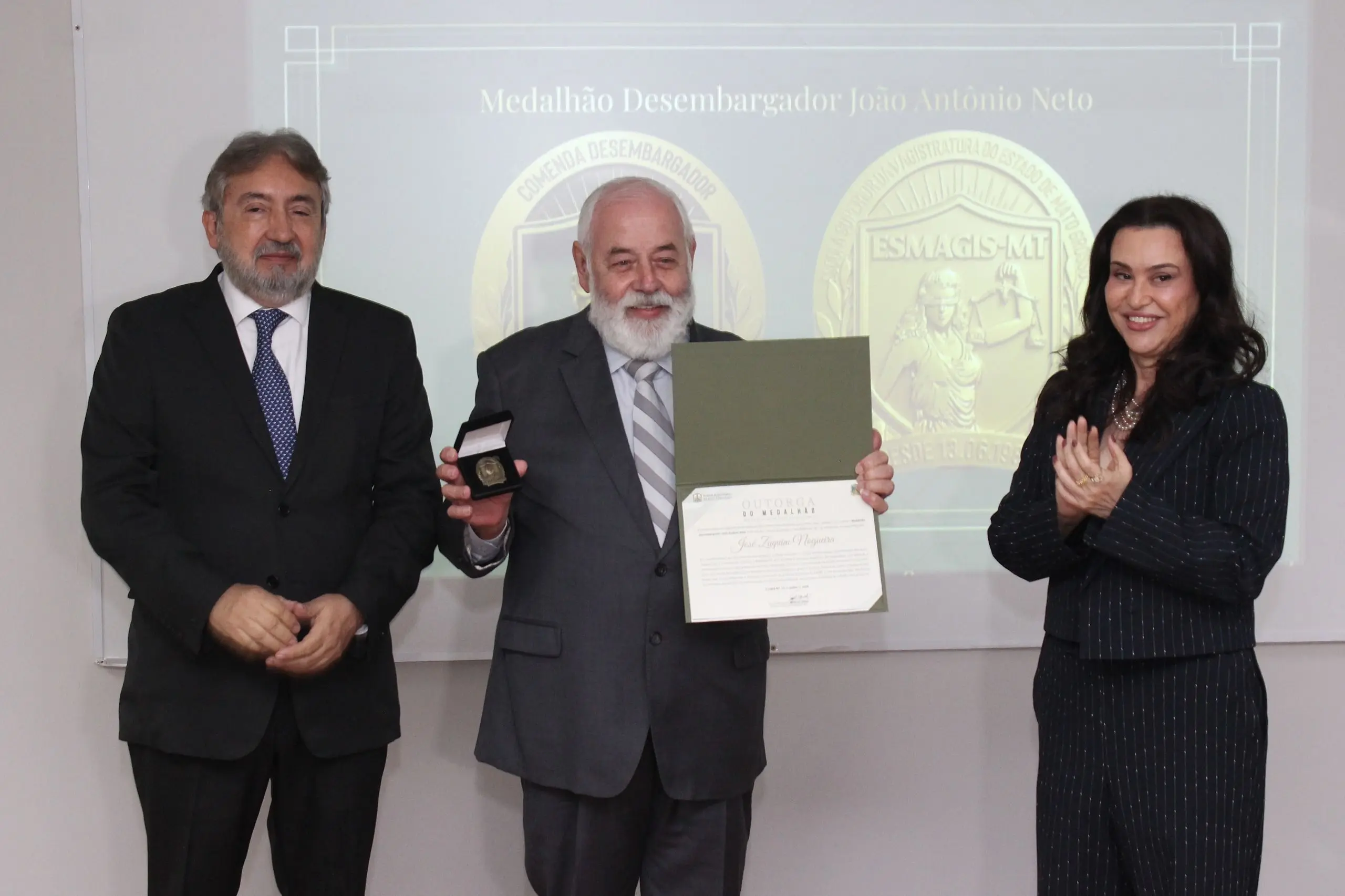 Homem de barba branca exibe medalha e diploma entre um homem de terno e uma mulher que aplaude. Ao fundo, projeção traz o título 