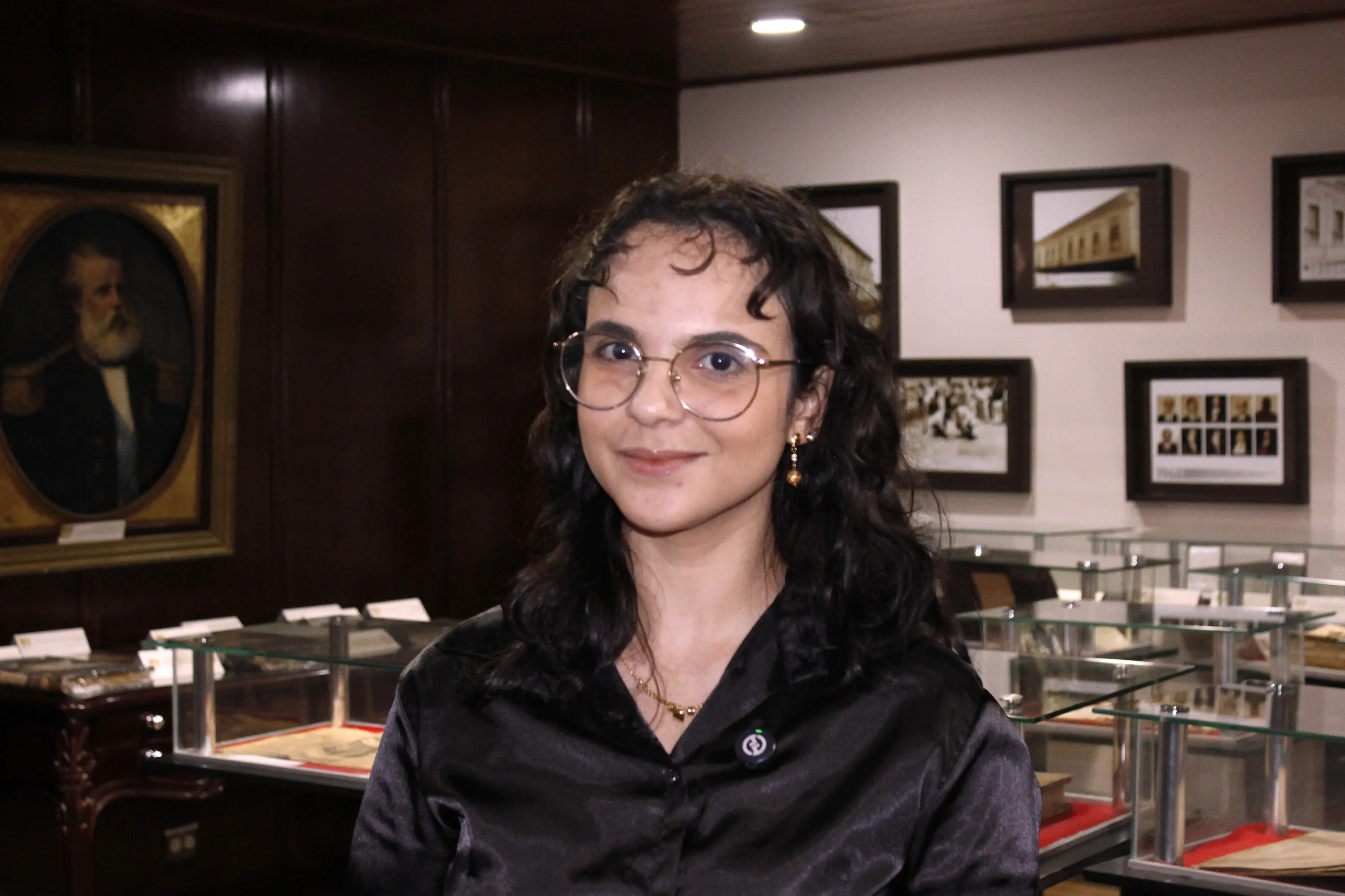 Mulher jovem de cabelos cacheados pretos, óculos redondos e blusa preta sorri. Ao fundo, quadros históricos e vitrines com documentos em exposição.