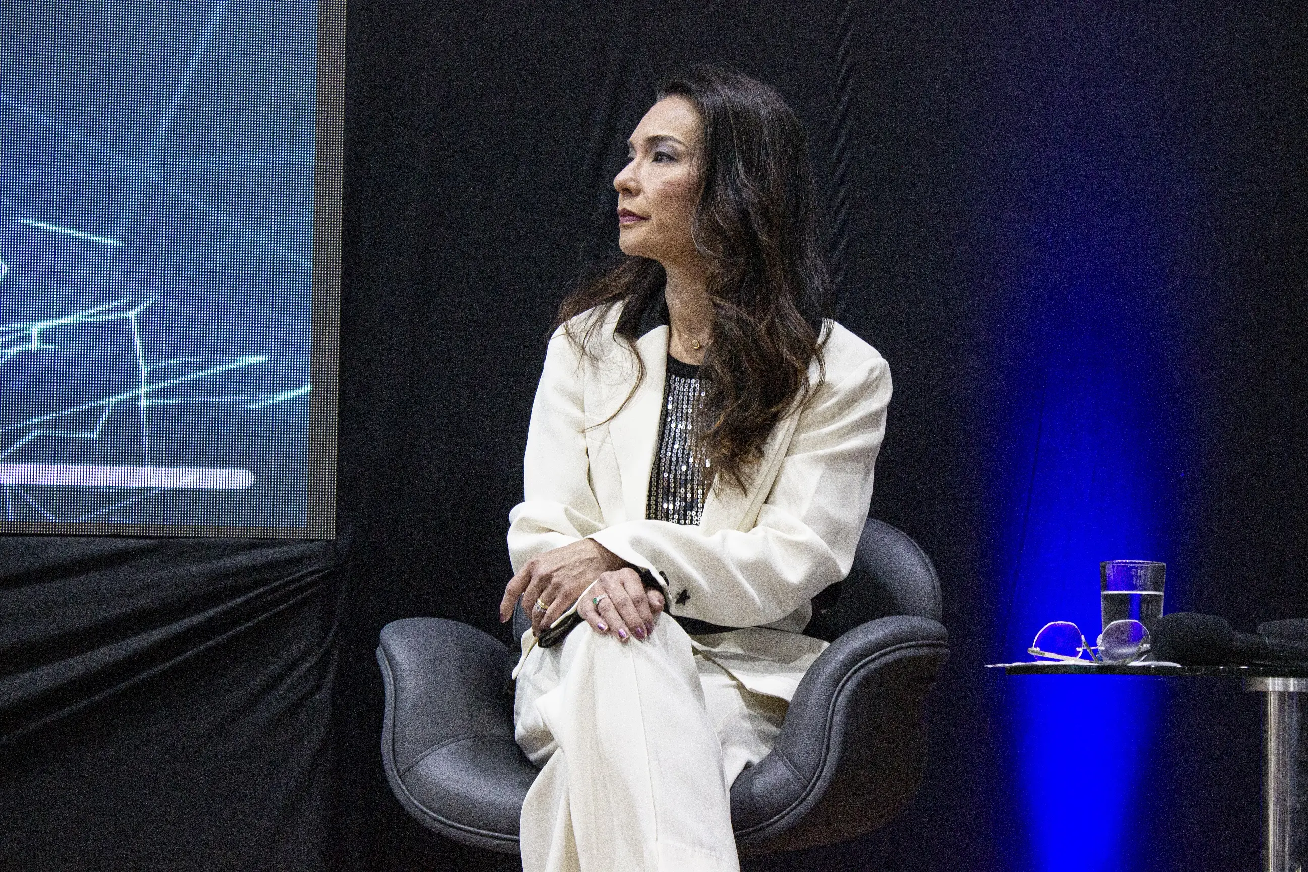 Mulher de cabelos longos e terninho branco sentada em uma poltrona escura de perfil, olhando para a esquerda. Ao lado, uma pequena mesa com óculos e um copo d'água sob fundo preto e azul.