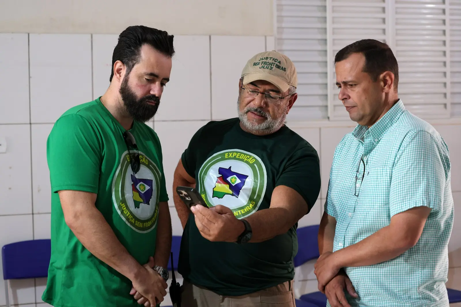 Juízes José Antônio Bezerra Filho e Flávio Fraga da Silva junto com o prefeito de Porto Esperidião, Odirlei Queiroz Faria estão em pé e conversam enquanto olham o celular. O do meio usa boné bege e camiseta verde-escura, homem da esquerda veste camiseta verde-clara e o da direita veste camisa xadrez