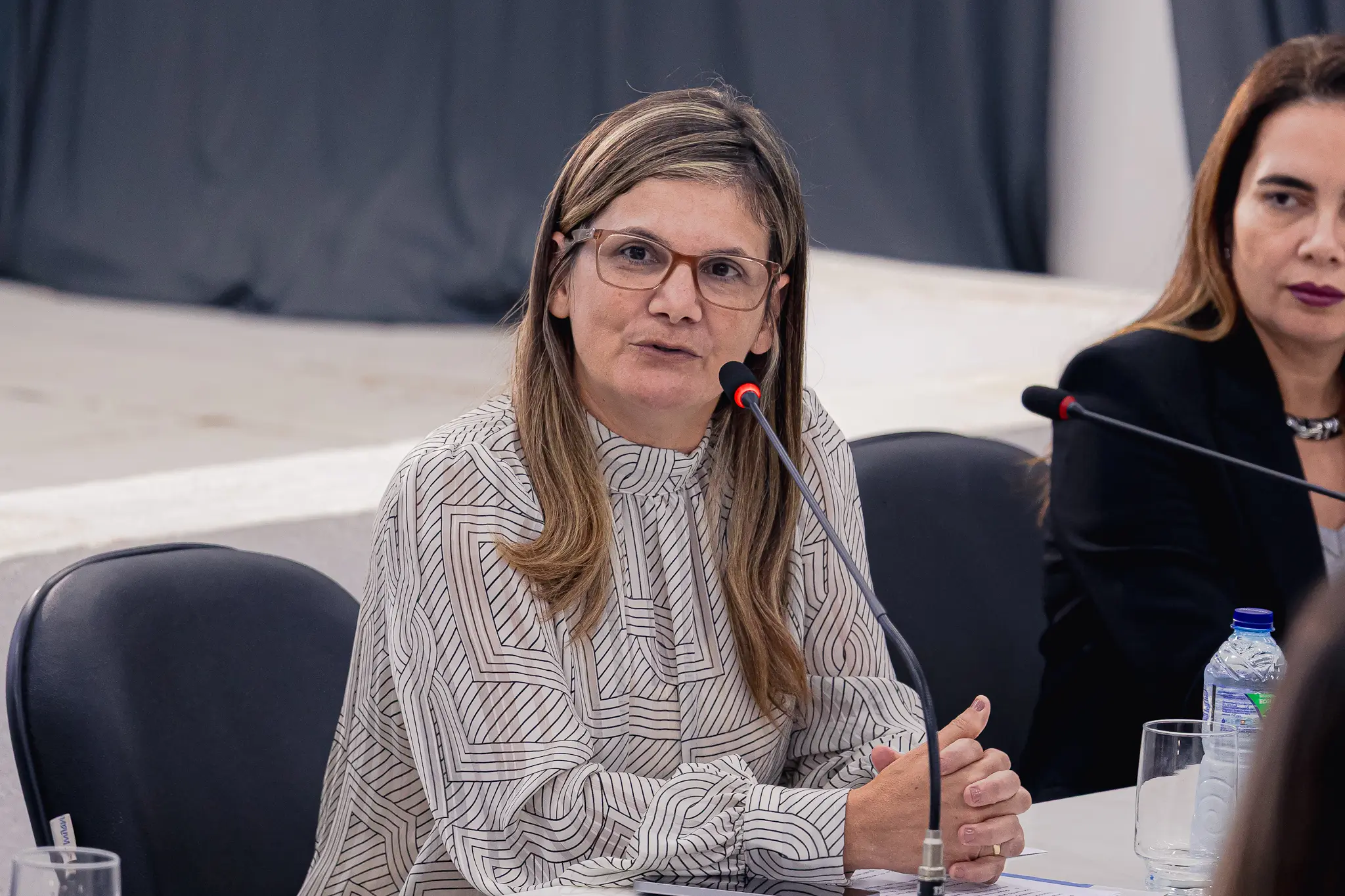 Uma mulher de óculos e blusa clara com estampas geométricas fala ao microfone, sentada à mesa com as mãos entrelaçadas. Ao lado direito, vê-se parte de outra mulher de blazer preto.