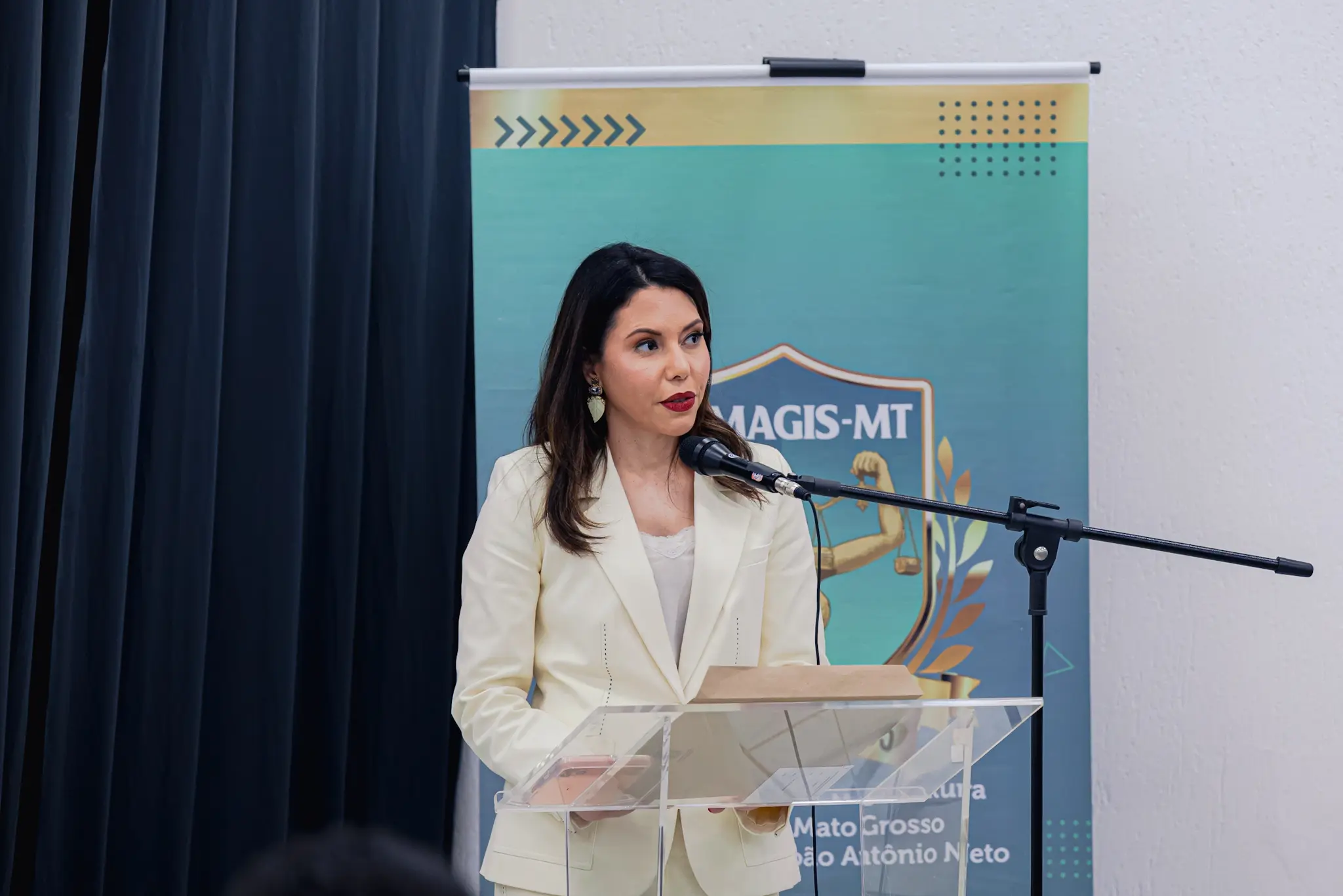 Uma mulher de cabelos escuros e blazer claro discursa em um púlpito acrílico com microfone. Ao fundo, uma cortina preta à esquerda e, à direita, um banner verde com o logotipo da MAGIS-MT.