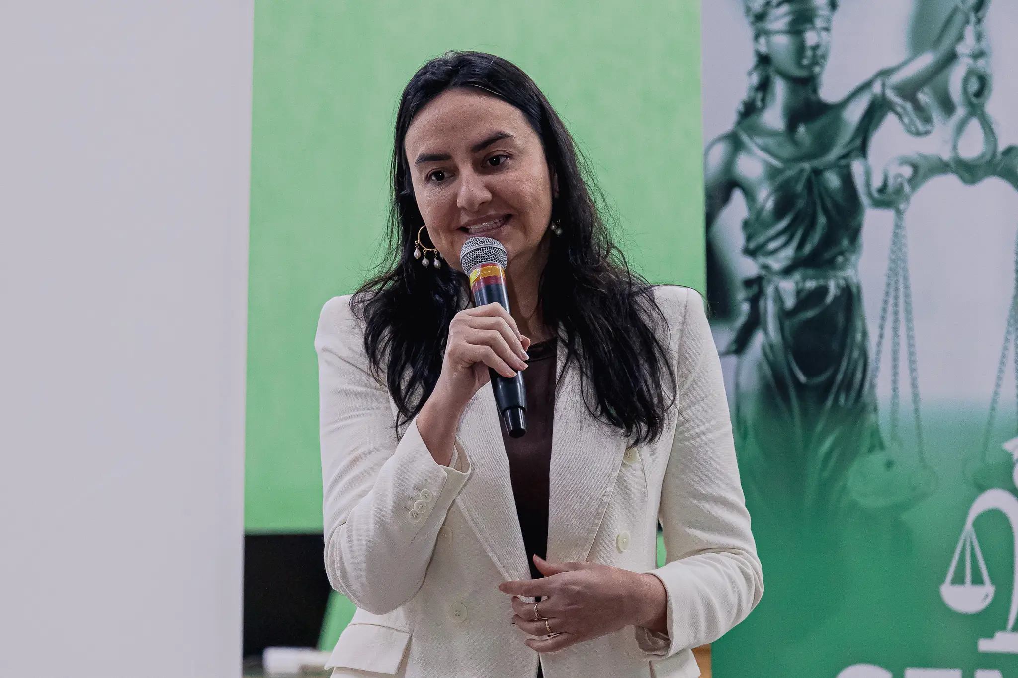 Mulher de cabelos longos e escuros fala ao microfone. Ela veste blazer off-white e brincos de argola. Ao fundo, um painel verde exibe a imagem da estátua da Justiça com a balança.