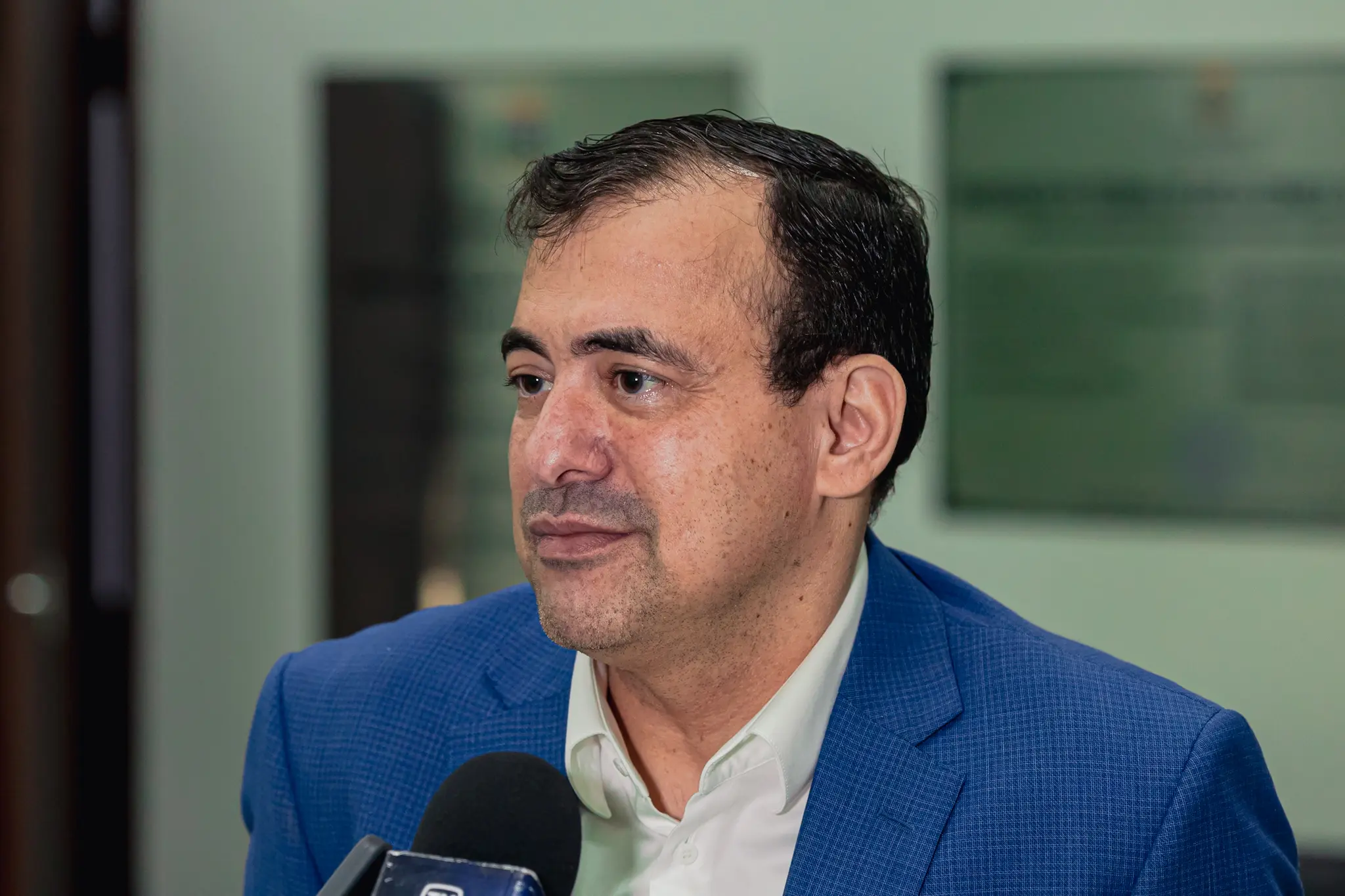 Homem de cabelos escuros e curtos, vestindo paletó azul e camisa branca, concede entrevista olhando para o lado esquerdo. Um microfone preto aparece em primeiro plano e o fundo está desfocado.