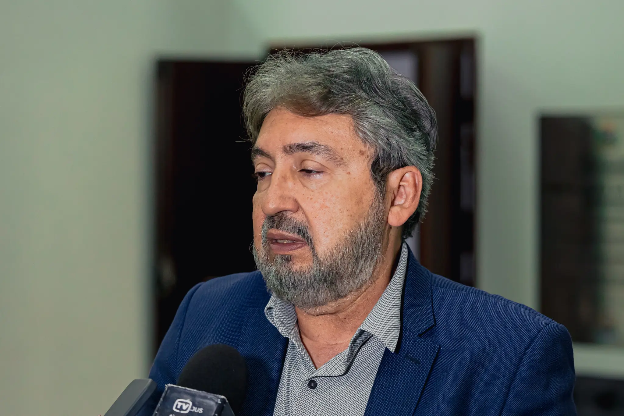 Homem com cabelos grisalhos e barba, fala direcionando o olhar para baixo e para a esquerda. Ele usa paletó azul e camisa social cinza. Um microfone da 