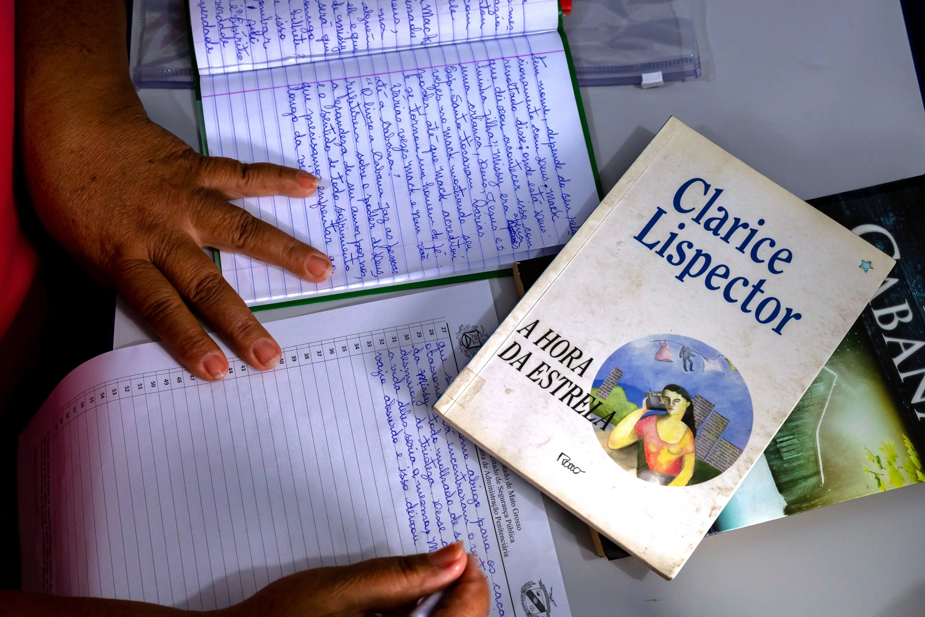 Sobre a mesa, um caderno aberto com anotações manuscritas é preenchido com atenção por uma pessoa. Ao lado, livros empilhados, incluindo obra de Clarice Lispector, indicam estudo e reflexão em ambiente de leitura orientada.