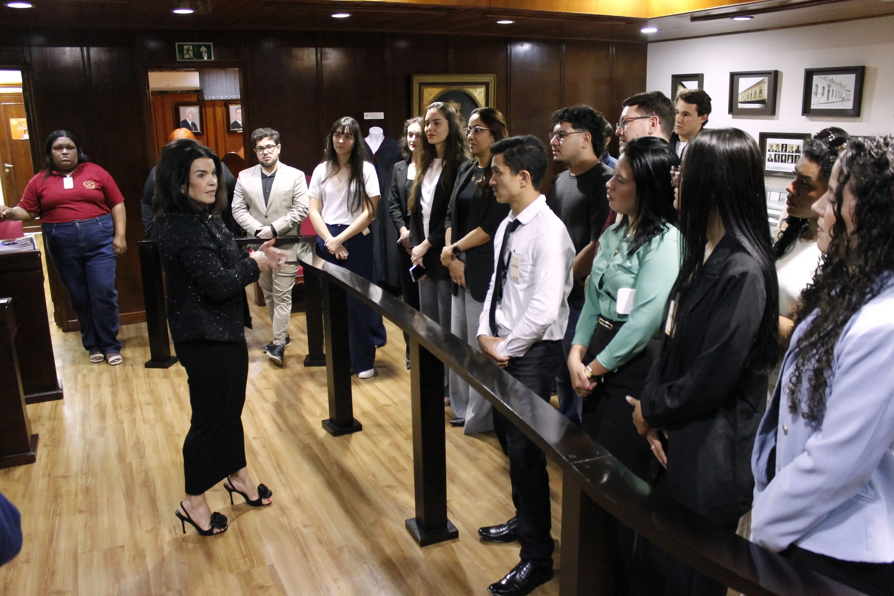 Mulher de roupa preta discursa para grupo de jovens em pé no Memorial do Judiciário Mato-Grossense, com fotos históricas ao fundo.
