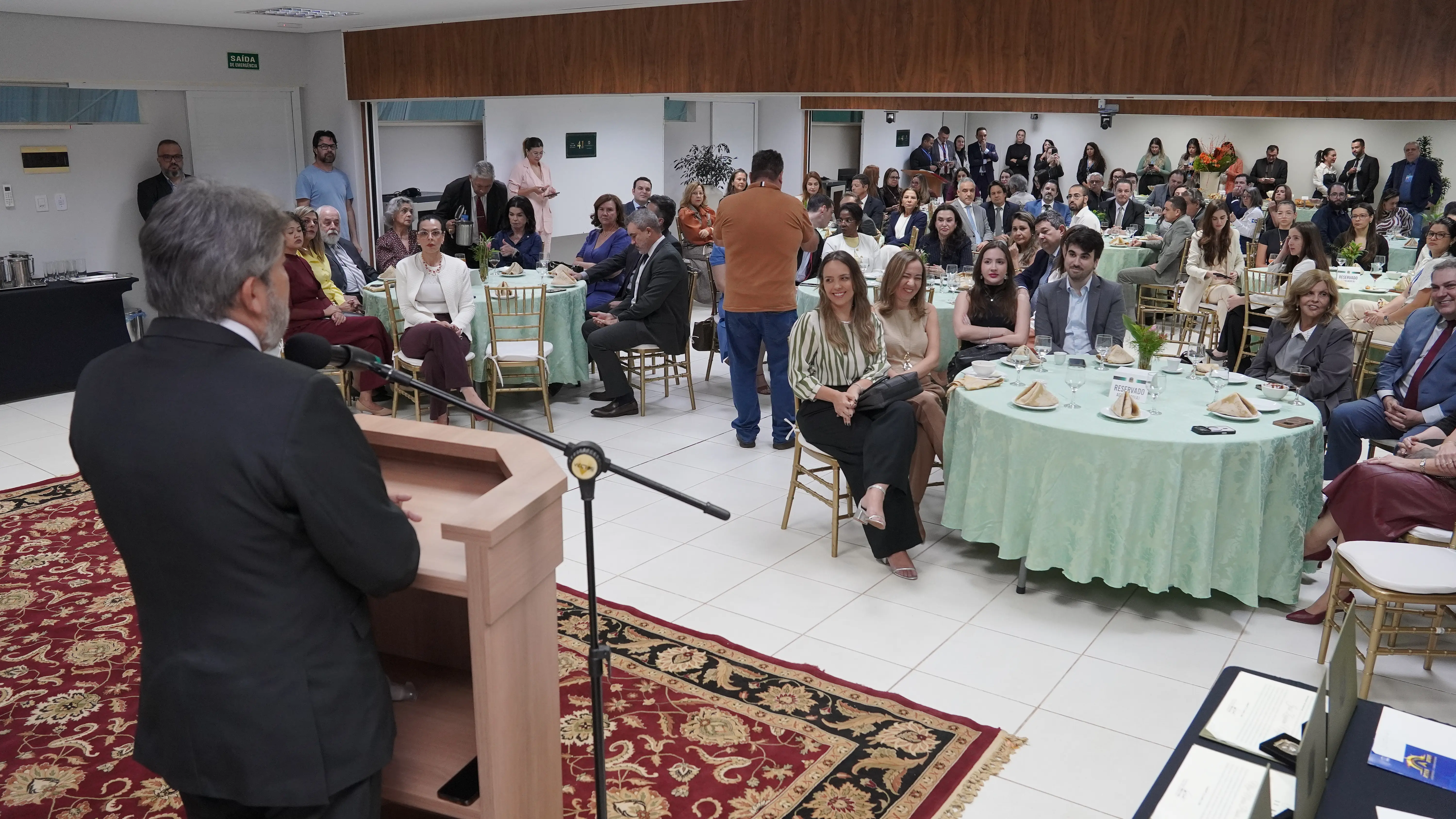 Ambiente interno amplo, com várias mesas redondas ocupadas por pessoas sentadas em cadeiras de madeira. Ao fundo e nas laterais, outras pessoas estão em pé, algumas registrando o momento com aparelhos. A disposição sugere um evento institucional ou social, com iluminação uniforme e paredes claras.