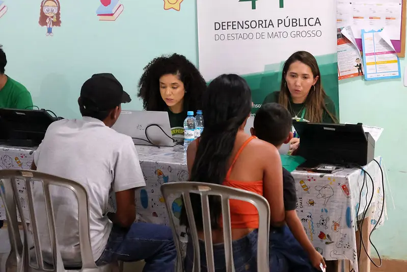Duas atendentes da Defensoria Pública de Mato Grosso, sentadas à mesa, conversam com um homem, uma mulher e uma criança, vistos de costas. Notebooks e garrafas de água estão sobre a mesa.