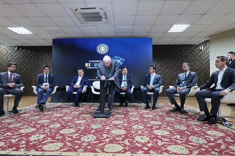 Foto horizontal que mostra o presidente do TJMT assinando a Resolução Conjunta nº 01/2025 durante a abertura da Conferência Recupera MT. Autoridades do sistema de justiça acompanham o ato em palco montado no auditório do Tribunal.
