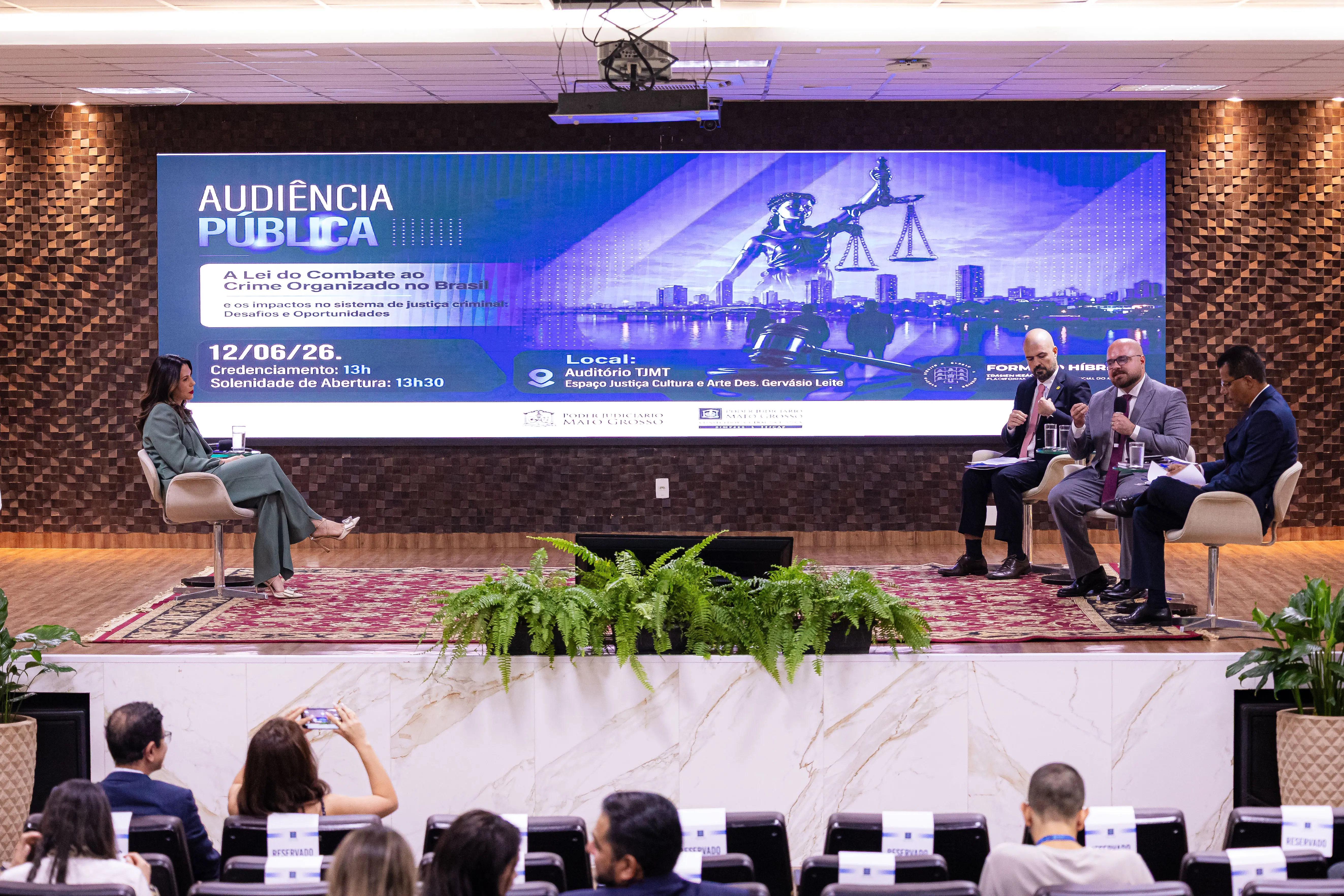Palco de auditório com telão azul ao fundo sobre audiência pública. Quatro pessoas estão sentadas em poltronas: uma mulher à esquerda e três homens à direita. Parte da plateia aparece abaixo.