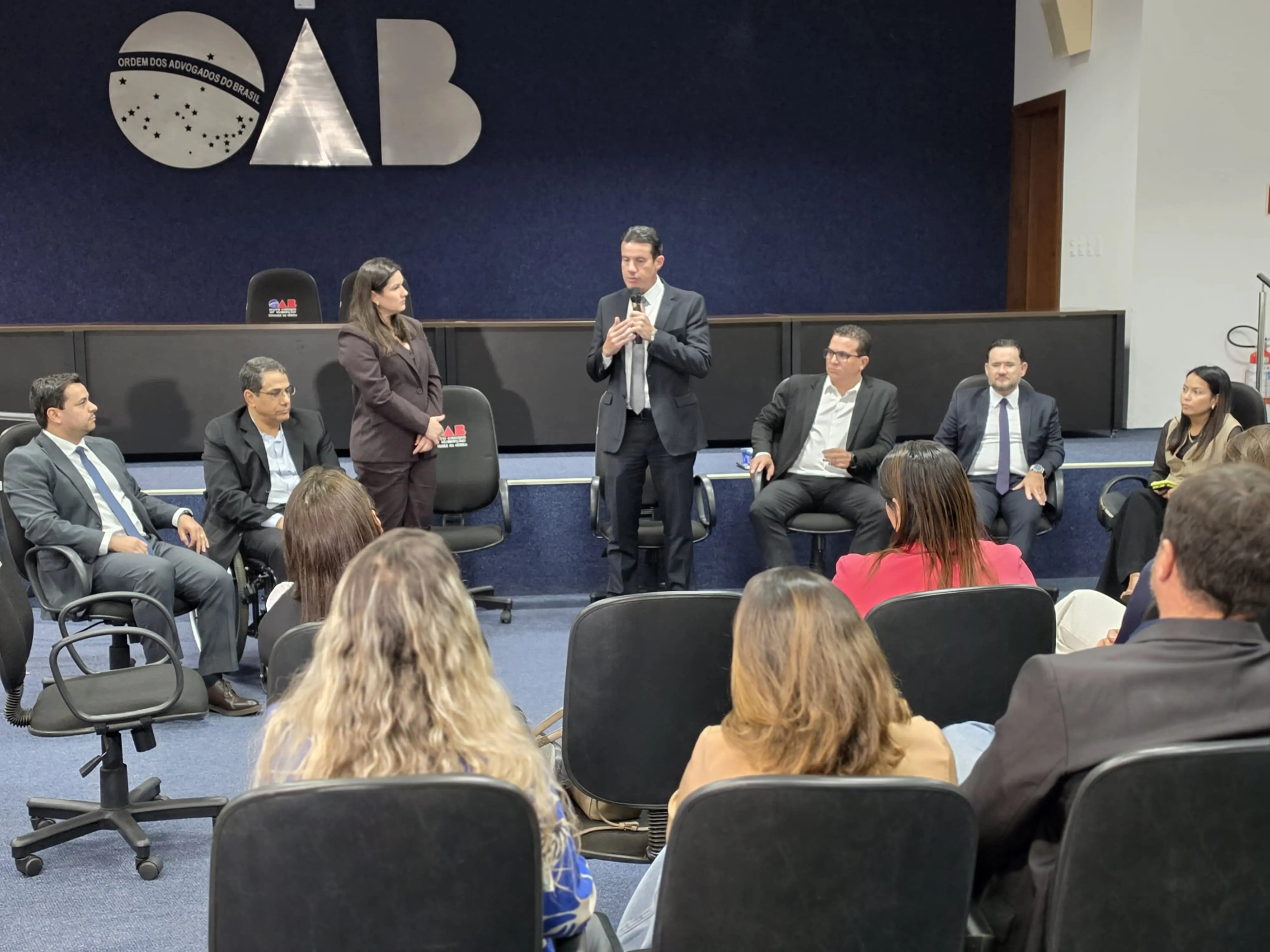 Foto horizontal que mostra várias pessoas sentadas em círculo e na plateia do auditório da OAB Tangará da Serra. Em pé, estão o desembargador Rodrigo Curvo e a advogada presidente daquela subseção da OAB. O local tem paredes azul marinho com o logotipo da OAB em metal prateado na parede ao fundo. 
