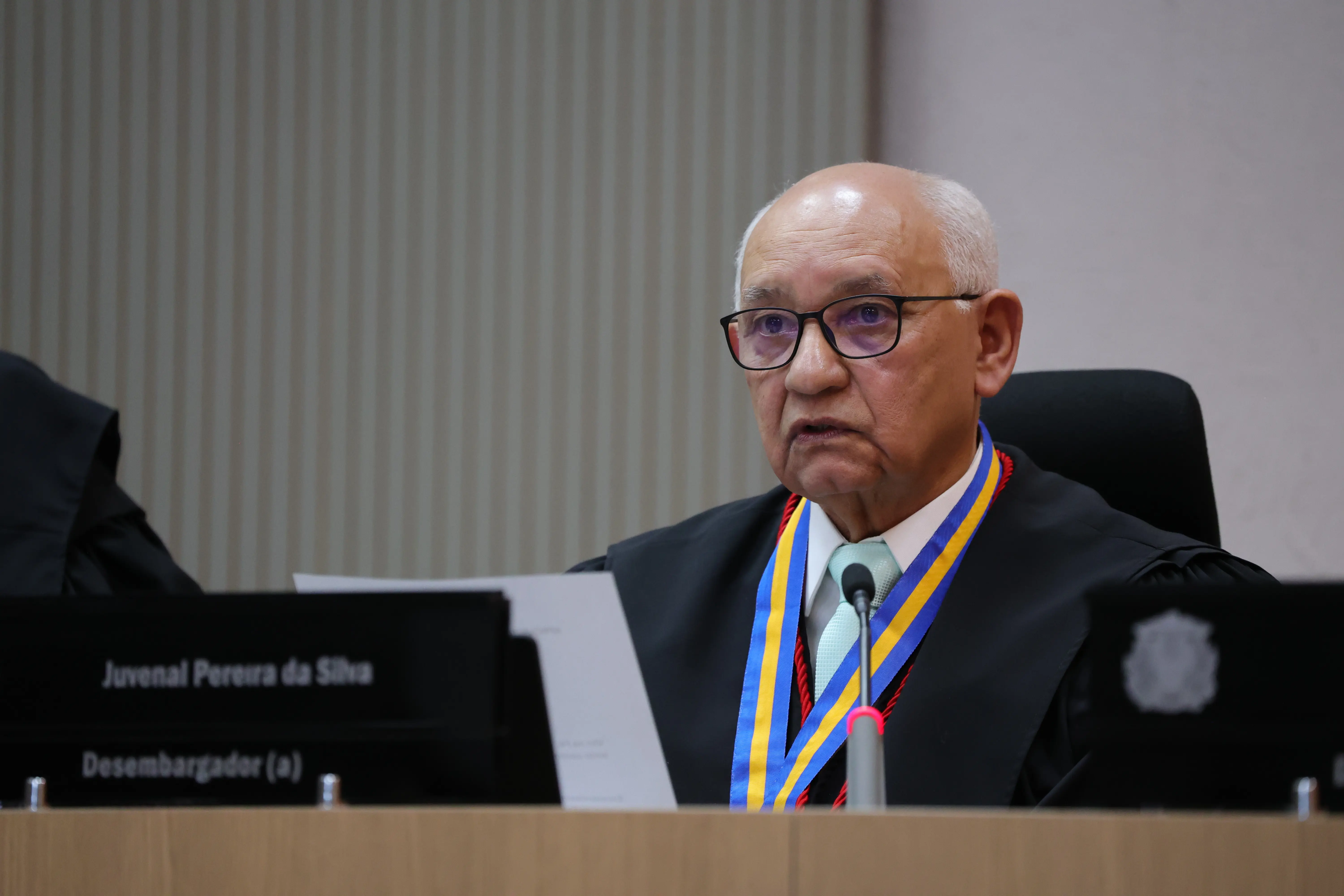 Desembargador Juvenal Pereira da Silva usa óculos, veste toga preta e colar com listras azuis e amarelas, segura folhas de papel branco. À sua frente, há uma placa preta de identificação onde se lê 