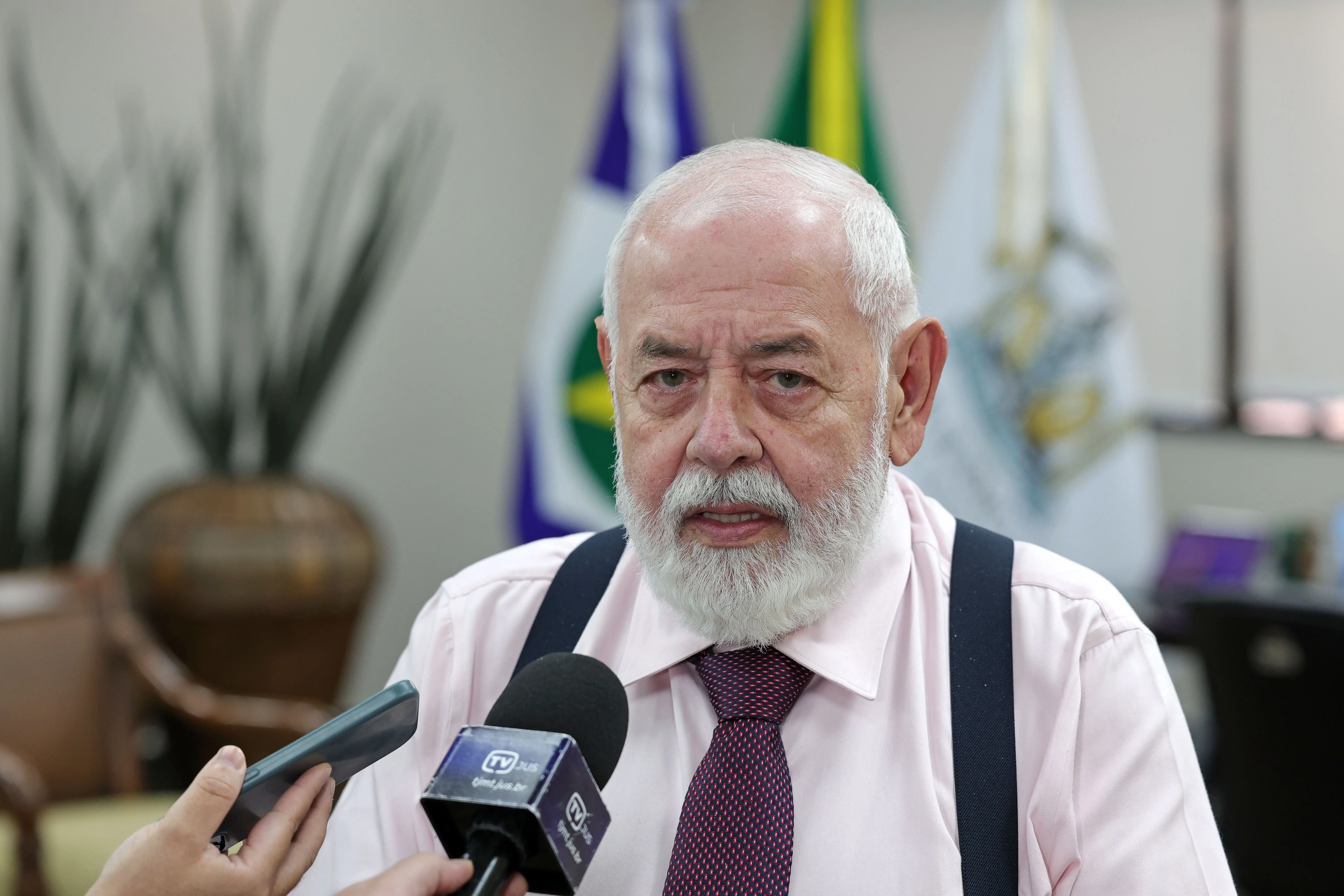 : Homem de barba branca, camisa rosa clara, gravata e suspensórios pretos concede entrevista. Um microfone da TV JUS e um celular são apontados para ele. Bandeiras ao fundo.