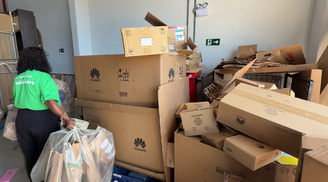 Imagem mostra uma sala com muitas caixas de papelão para reciclagem. Uma mulher aparece de costas, segurando sacos plásticos transparentes cheios de papelão dobrado. 