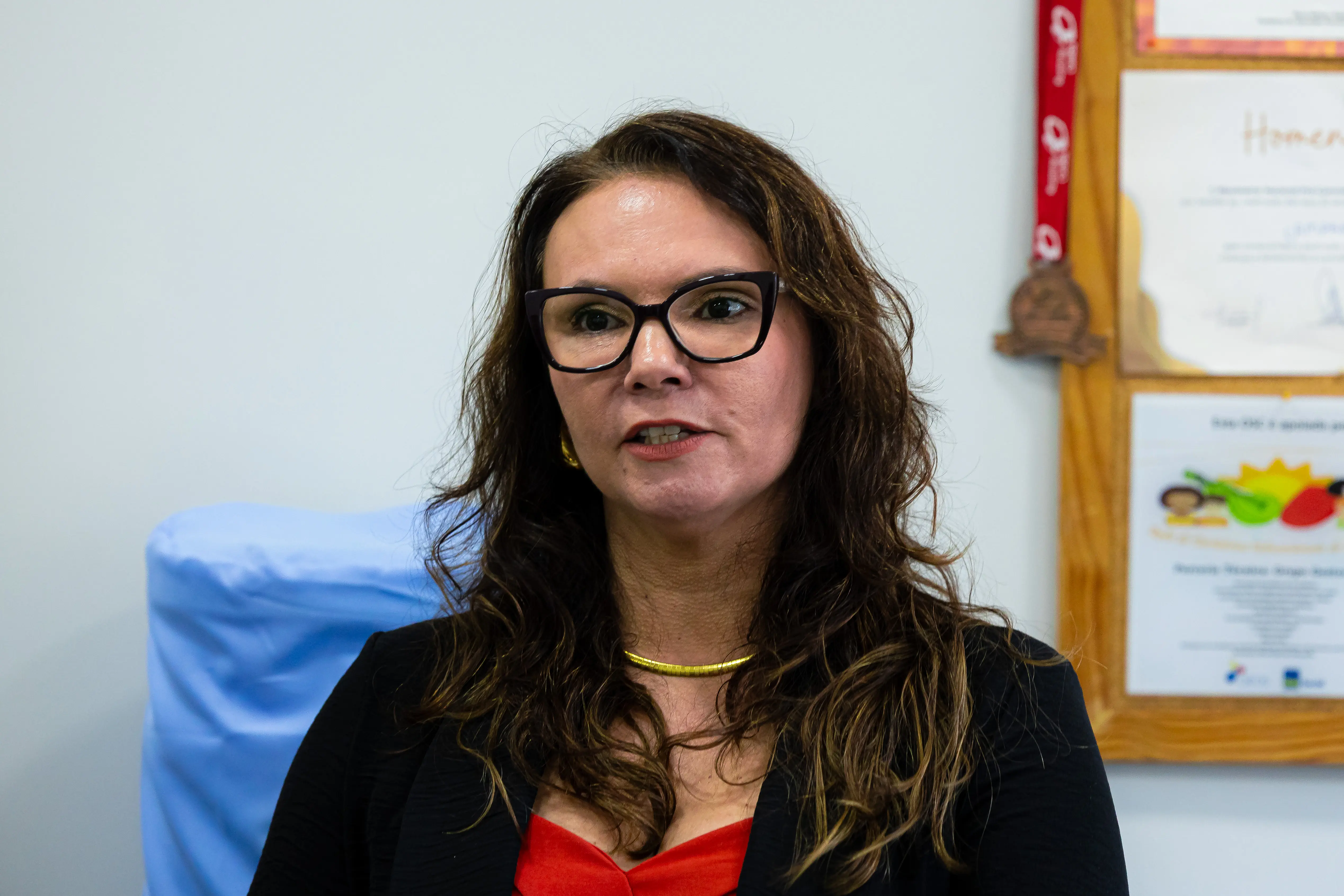 Mulher de cabelos castanhos cacheados e óculos de armação escura fala em ambiente interno. Veste blazer preto sobre blusa vermelha. Ao fundo, quadro com certificado emoldurado.