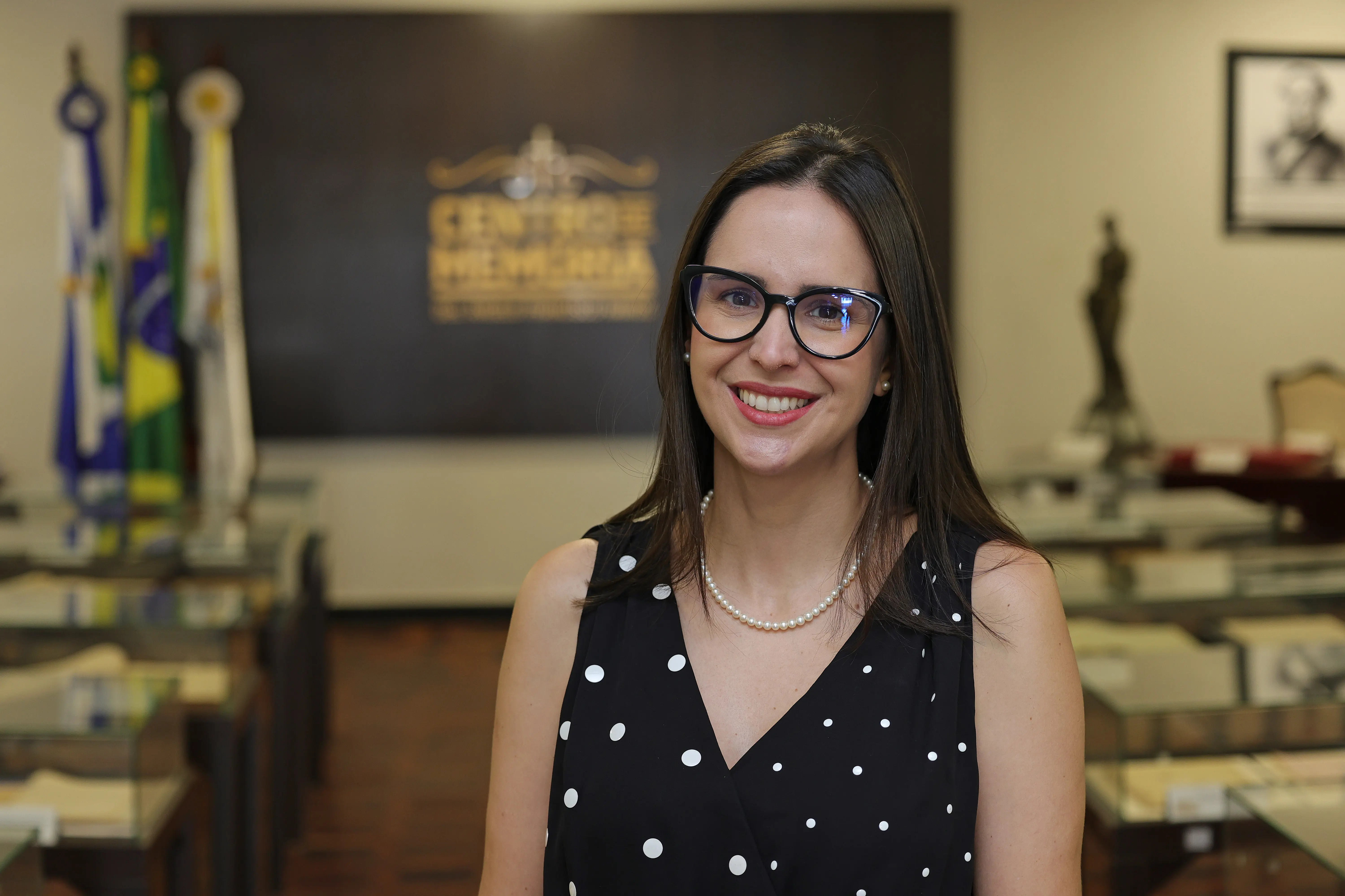 Coordenadora administrativa do TJMT, Bruna Penachioni é uma mulher sorridente de pele clara, cabelos escuros, longos e liso, usa óculos de grau pretos e colar de pérolas. Veste blusa preta com bolinhas brancas. Ao fundo desfocado, há uma placa escura com letras douradas, bandeiras e balcões de vidro