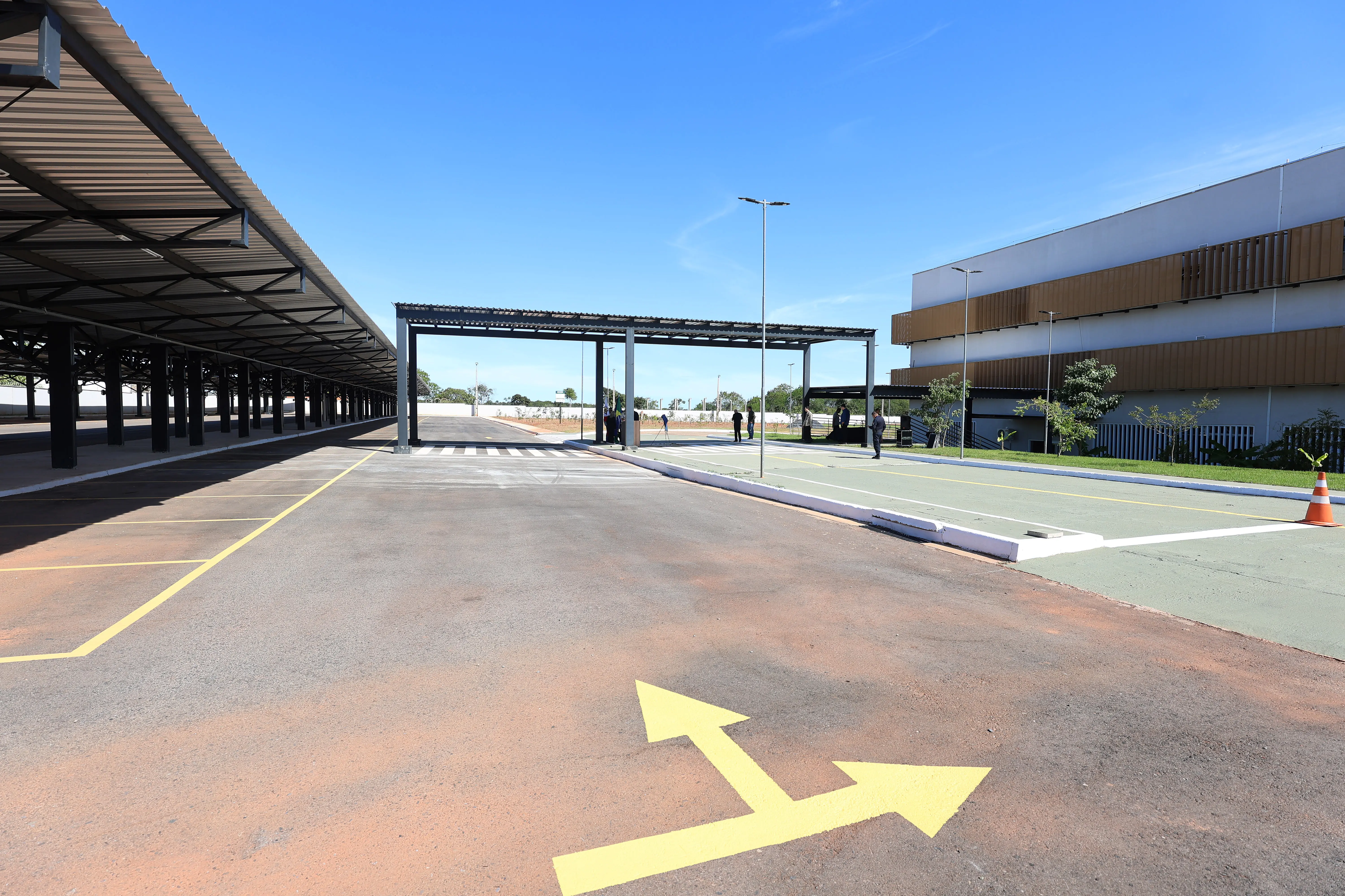 Vista ampla de um estacionamento aberto sob céu azul. À esquerda, vagas cobertas por estrutura metálica; à direita, um prédio moderno. No asfalto, em primeiro plano, uma seta amarela indica direções.