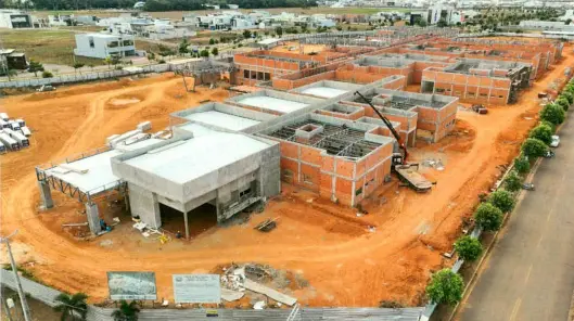 Vista aérea das obras do Fórum de Sinop. Um grande complexo em construção com estruturas de concreto e paredes de tijolos vermelhos expostos, cercado por terra batida e uma avenida arborizada.