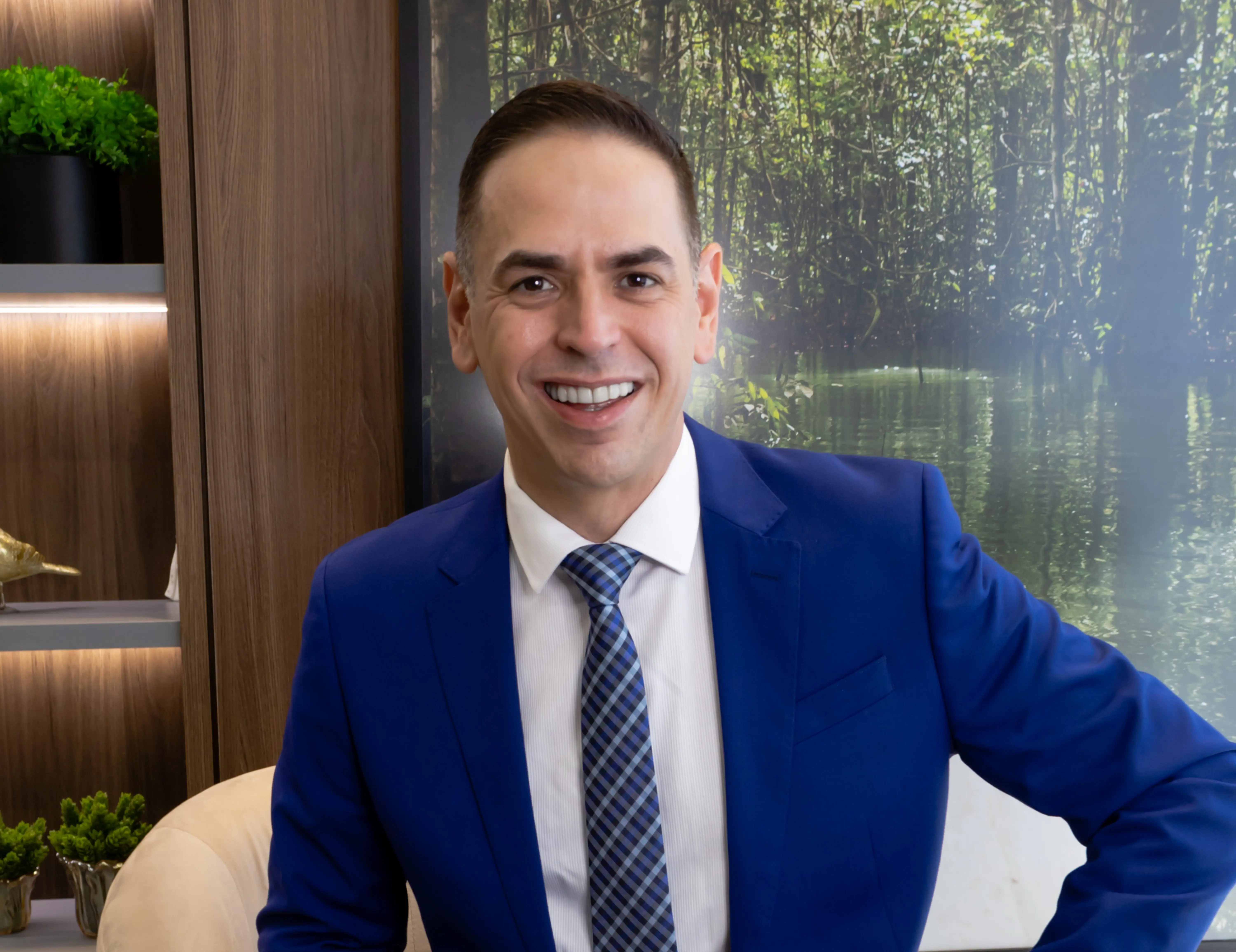 O professor Carlos aparece, de frente e sorrindo, em meio corpo. Veste terno, camisa branca e gravata estampada. No fundo, há um painel de madeira com prateleiras e vasos de plantas