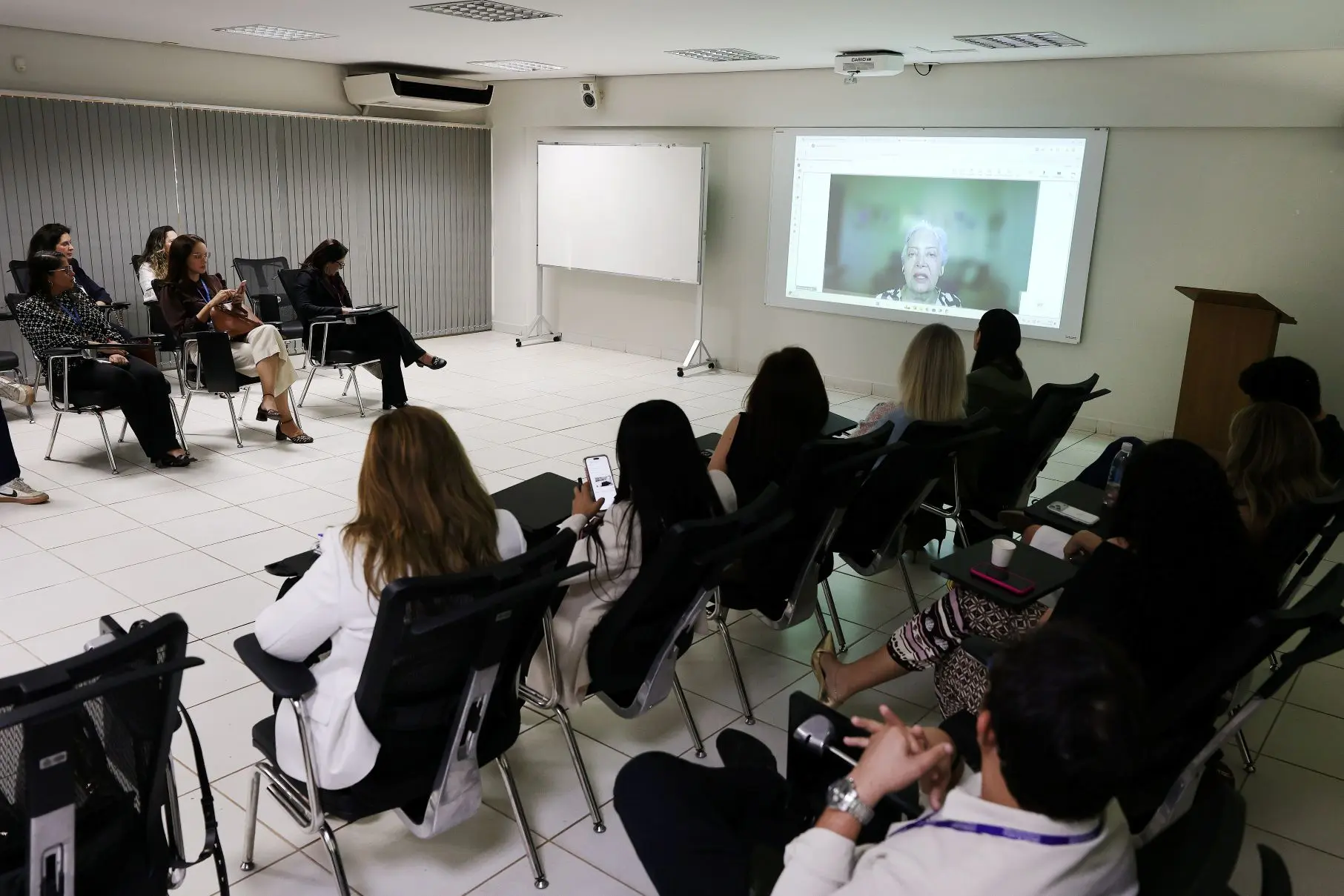 Foto horizontal que mostra uma sala de aula com diversas pessoas sentadas em carteiras e olhando para o telão, onde se projeta a imagem da desembargadora Clarice Claudino, que participou por videoconferência da apresentação da ferramenta ClarIA.