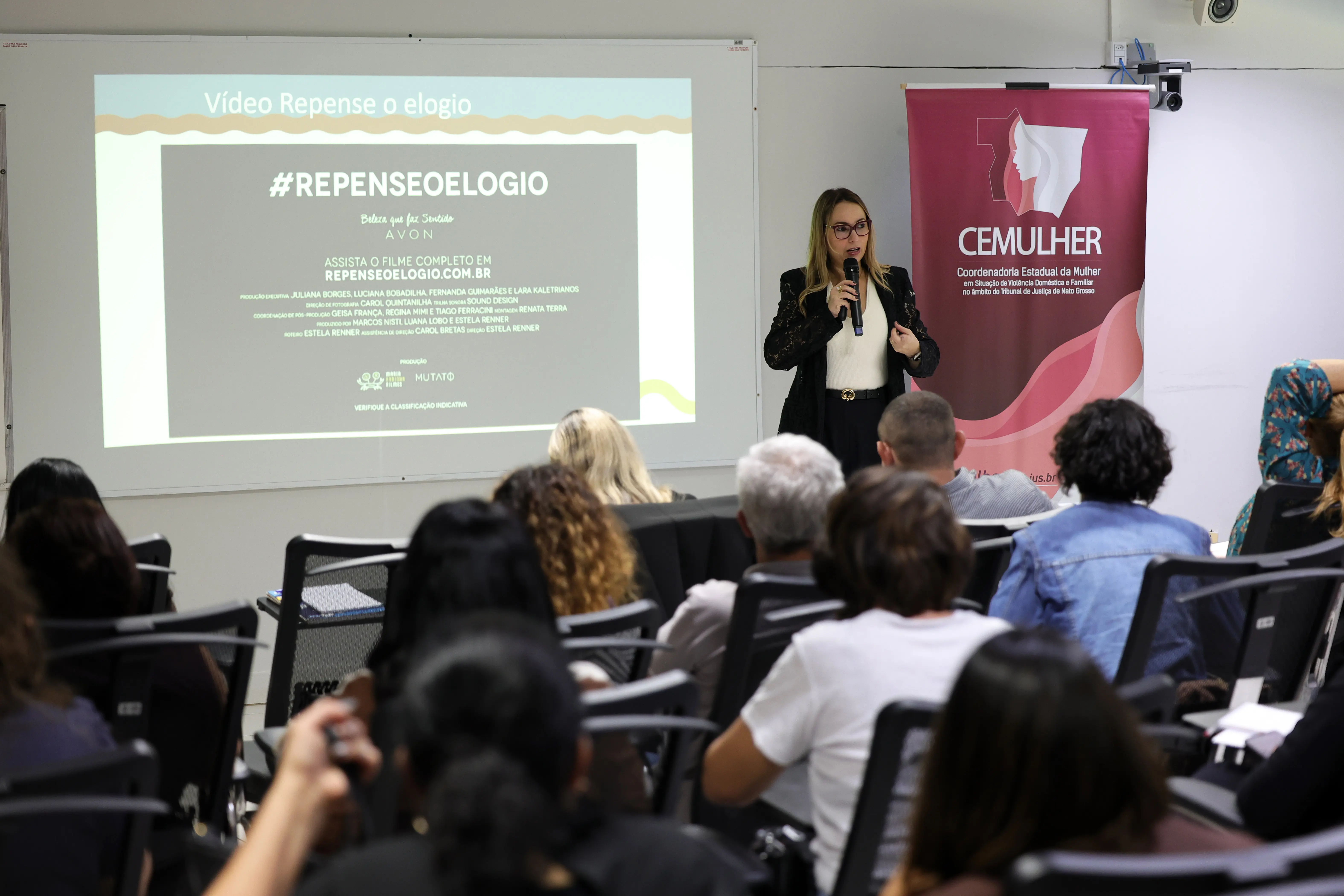 Mulher de blazer preto fala ao microfone diante de plateia sentada. Ao fundo, telão com slide sobre campanha e banner do CEMULHER - Coordenadoria Estadual da Mulher