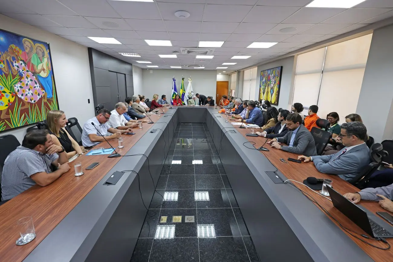 Foto horizontal que mostra a sala de reuniões da Presidência do TJMT lotada de pessoas sentadas em volta da mesa em formato de U.