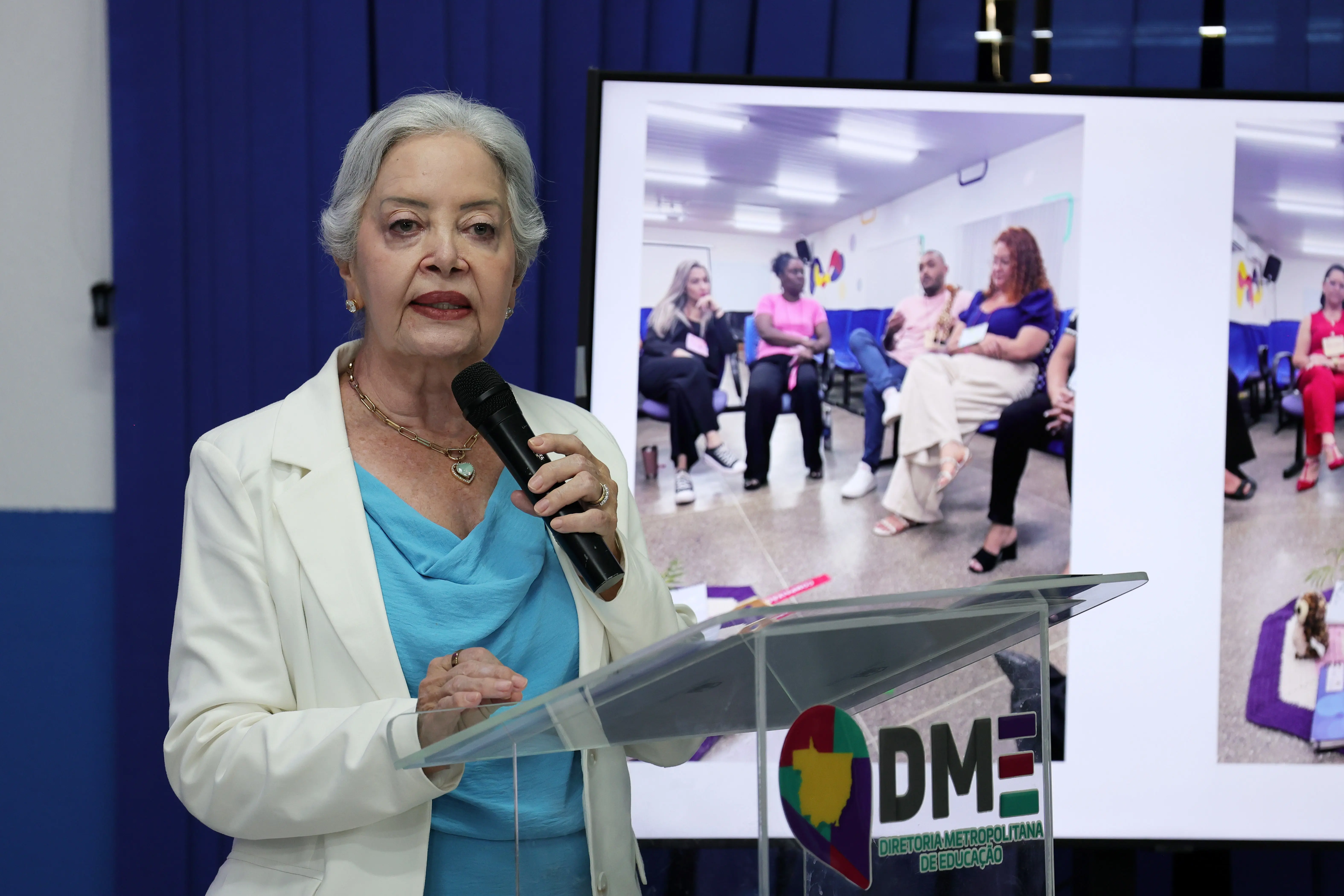 Mulher de cabelos brancos, blazer branco e blusa azul-turquesa, fala ao microfone em púlpito com o logotipo da Diretoria Metropolitana de Educação. Ao fundo, telão exibe foto de participantes reunidos em roda de conversa.