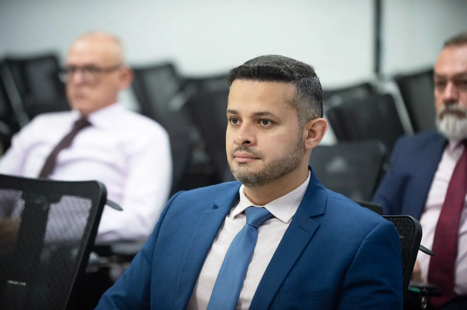 Homem de terno azul e barba por fazer sentado em plateia. Ao fundo, dois homens mais velhos de camisa e gravata, desfocados. 