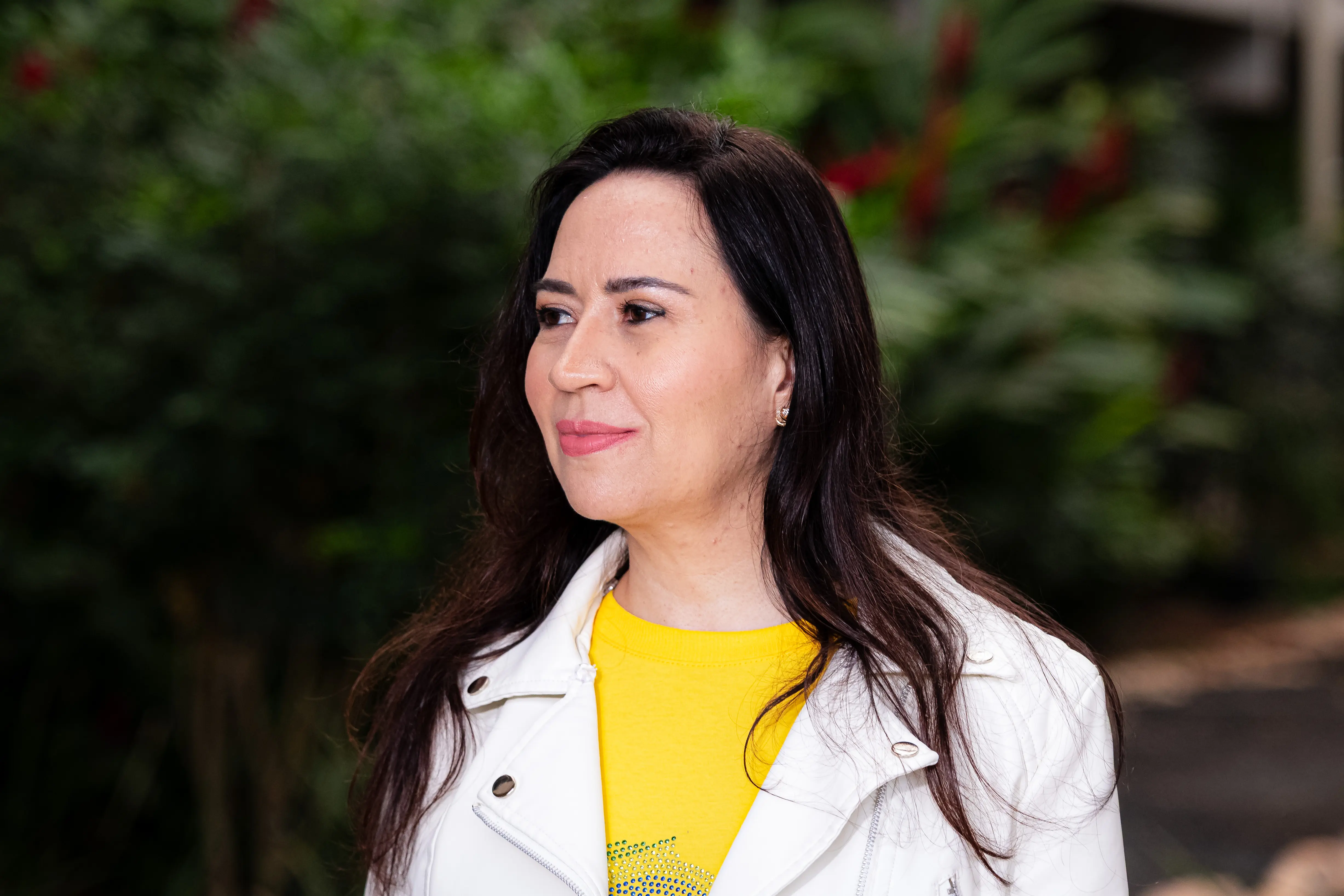 Mulher de cabelos longos e escuros veste jaqueta branca sobre blusa amarela com detalhes em azul, semblante alegre, olha para o lado. Ao fundo, vegetação desfocada